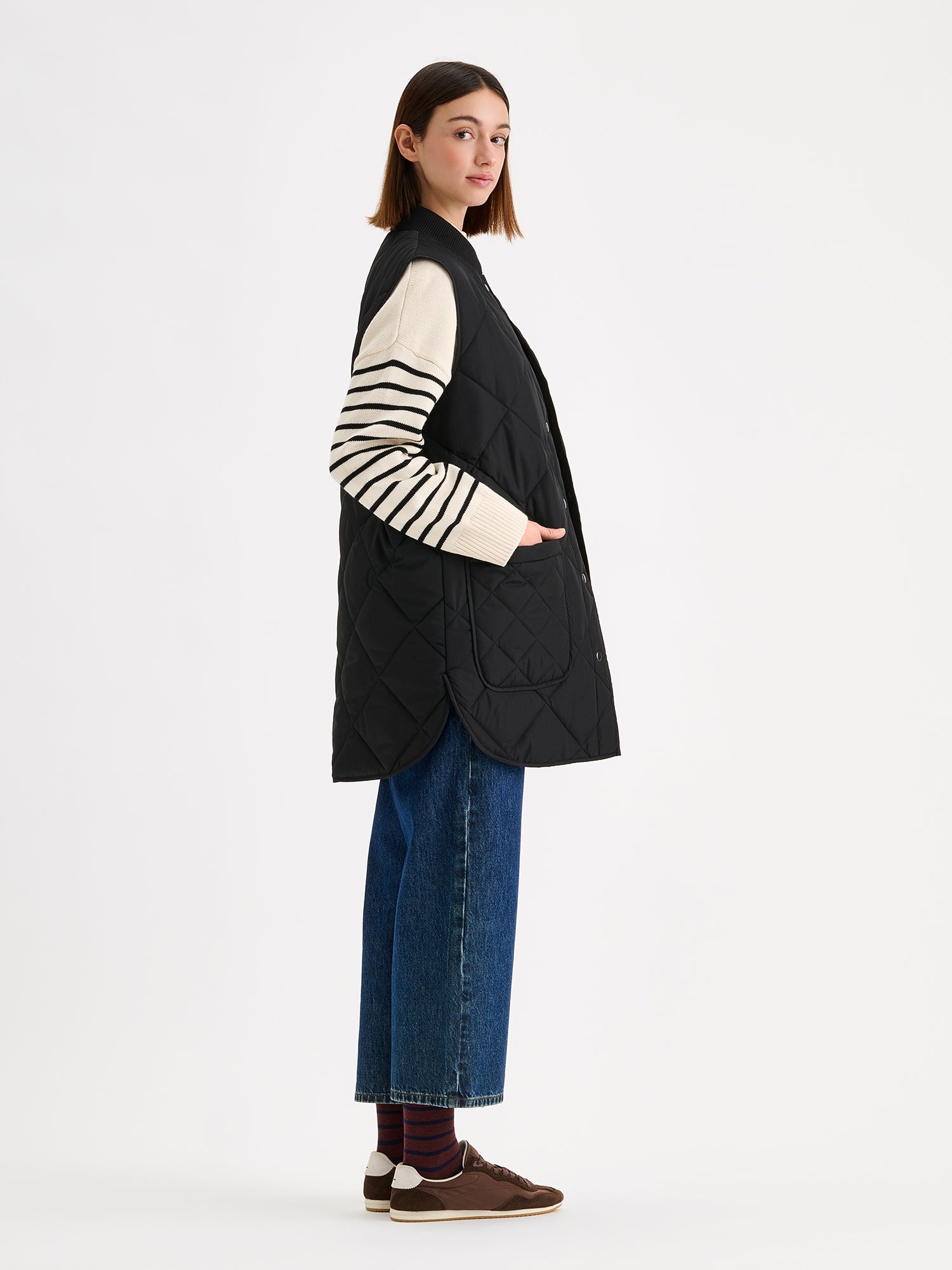 Nanso women's beautiful classic quilted vest in black, on a model with a striped sweater and wide blue jeans, from the side.