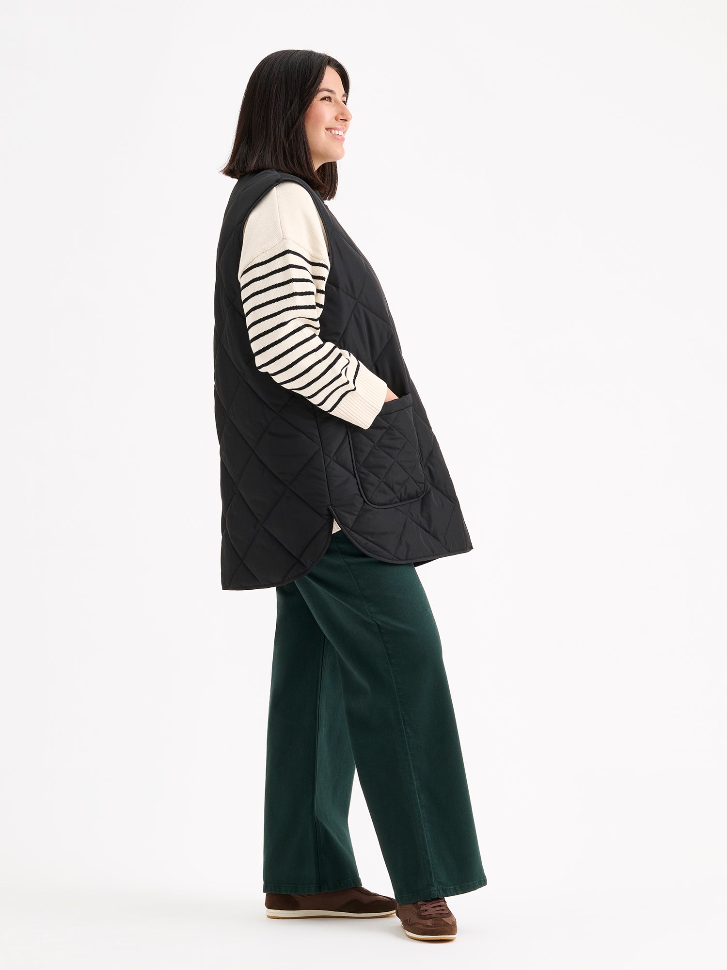 Nanso women's beautiful classic quilted vest in black, on a model with a striped sweater and wide blue jeans, side view.