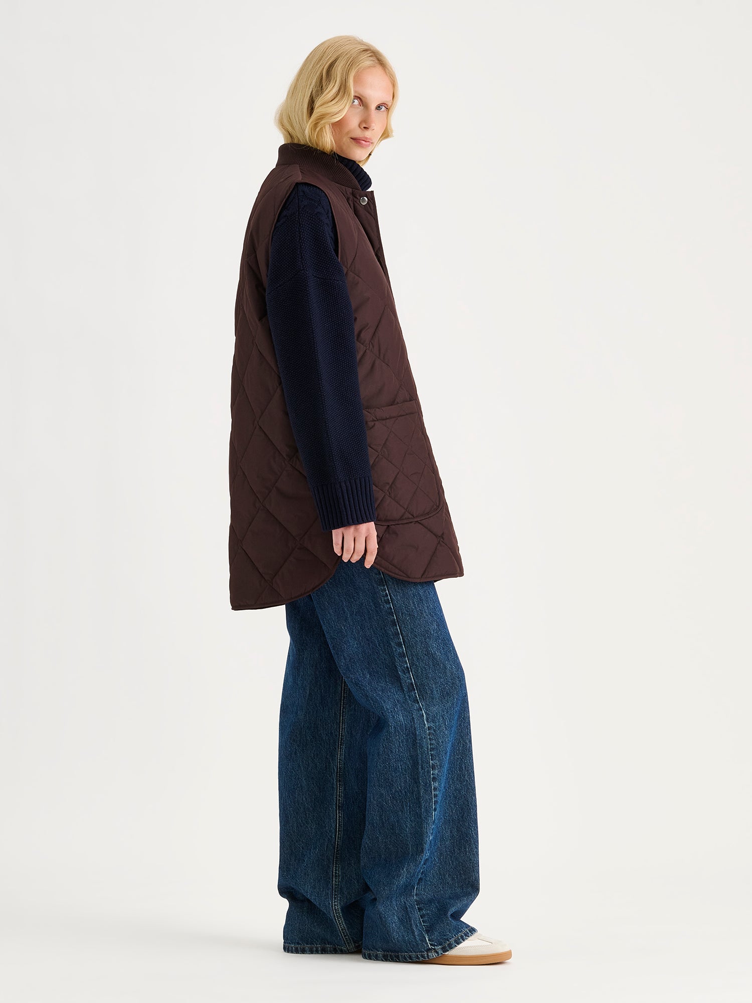 Nanso women's beautiful classic quilted vest in dark brown on a blonde model with a sweater and blue jeans, side angle.