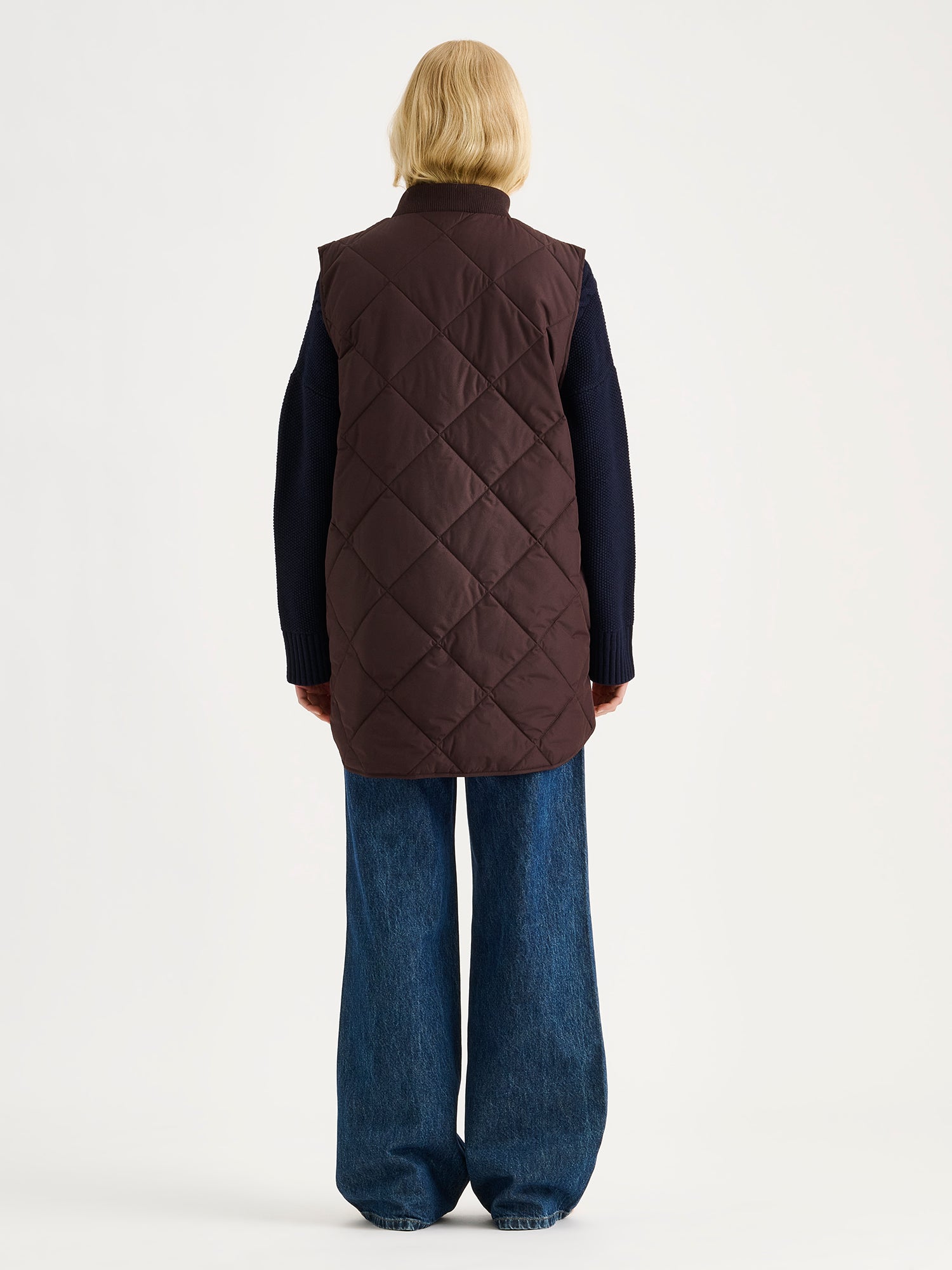 Nanso women's beautiful classic quilted vest in dark brown on a blonde model with a sweater and blue jeans, from the backside.