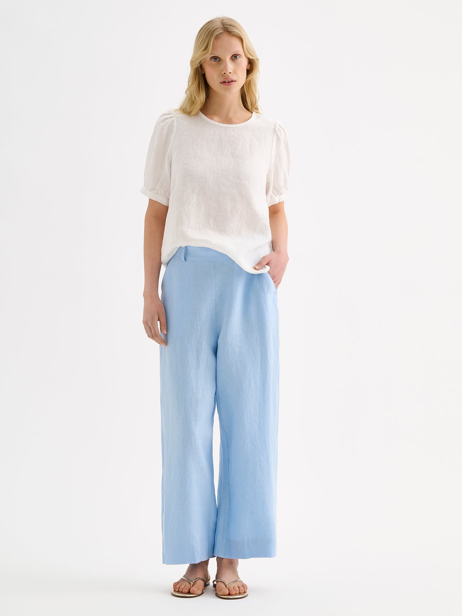 Nanso women's white breathable Sarastus linen blouse featuring short puffed sleeves on a model with light blue linen trousers and sandals.