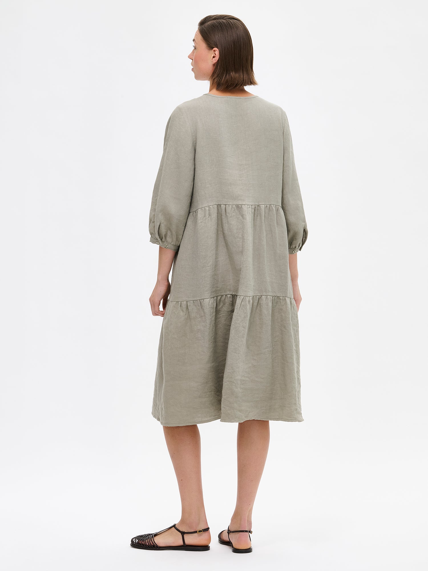 Nanso women's SARALIVIA linen dress in green features charming layered ruffles, 3/4 sleeves, side pockets and a V-neckline. Pictured on a model with sandals on from the backside.
