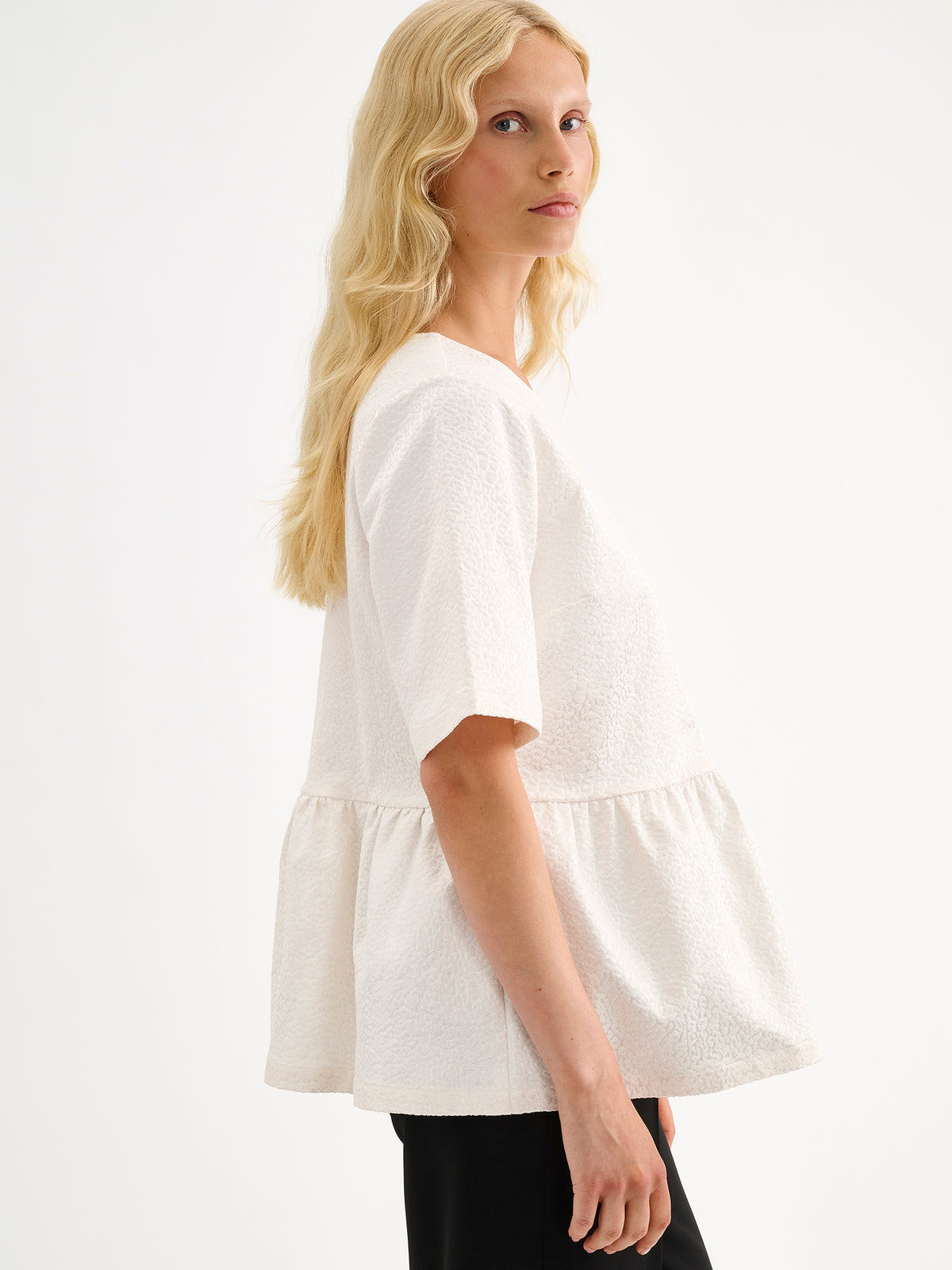 Nanso women's beautiful and elegant natural white Leora blouse captivates with its peplum-style silhouette and striking fabric, combining a subtle sheen with a richly textured surface. Pictured on a model with black trousers.
