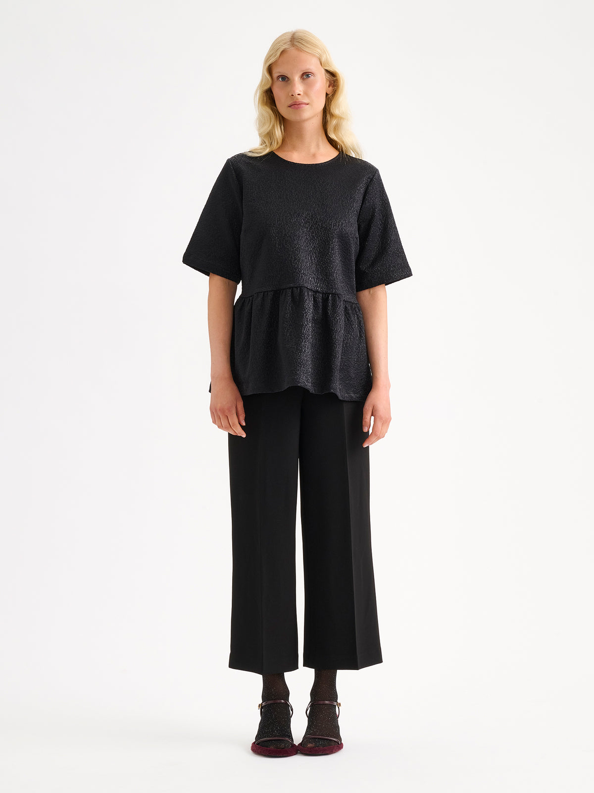 Nanso women's beautiful and elegant black Leora blouse captivates with its peplum-style silhouette and striking fabric, combining a subtle sheen with a richly textured surface, on a model with black trousers. | 36 - 174cm