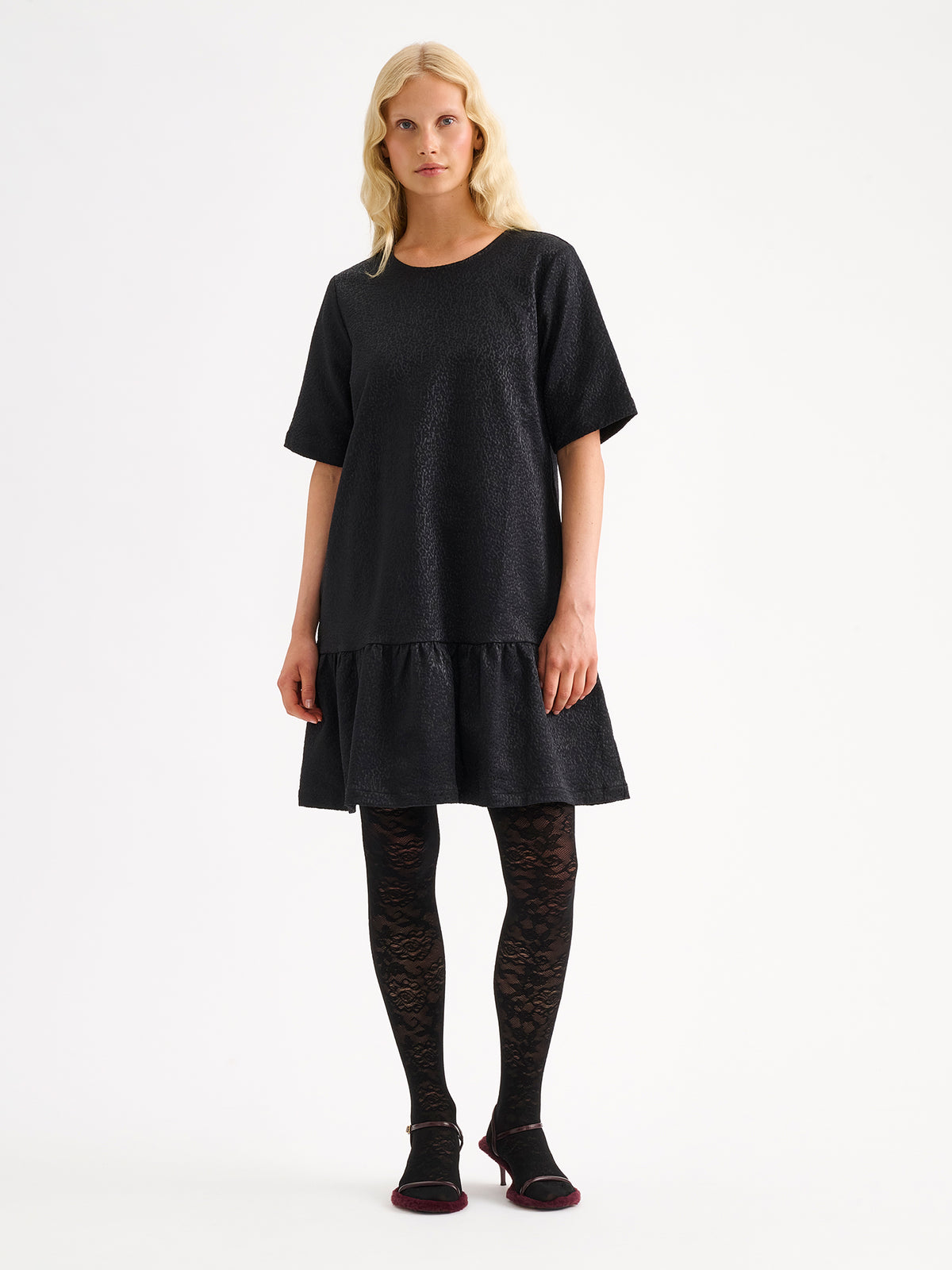 Nanso women's striking black Leora dress captivates with its beautiful fabric, combining a festive sheen with a rich textured surface, on a model with heels. | 36 - 174cm