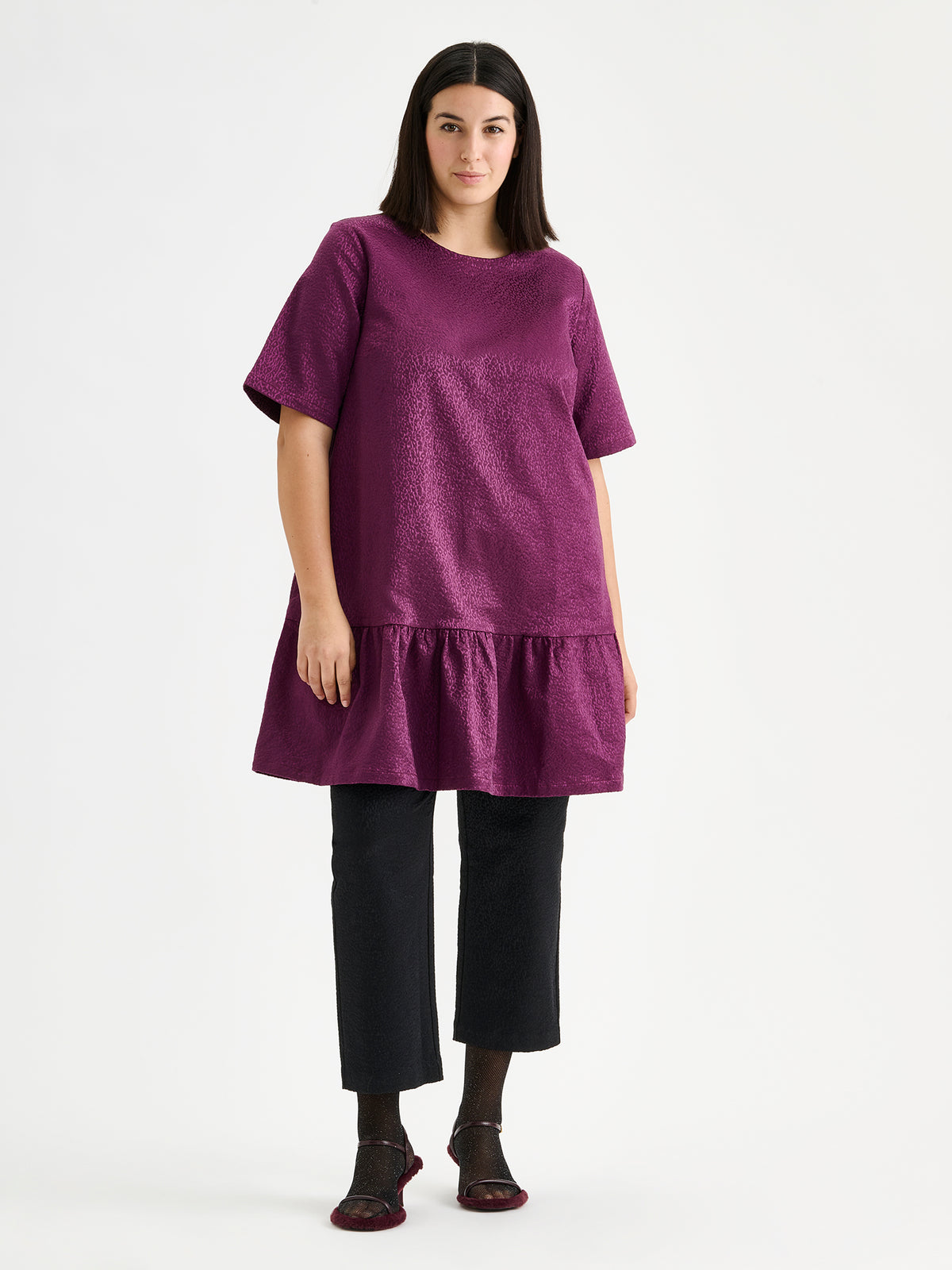 Nanso women's striking Leora dress captivates with its beautiful fabric, combining a festive sheen with a rich textured surface, pictured on a model with heels and black trousers.