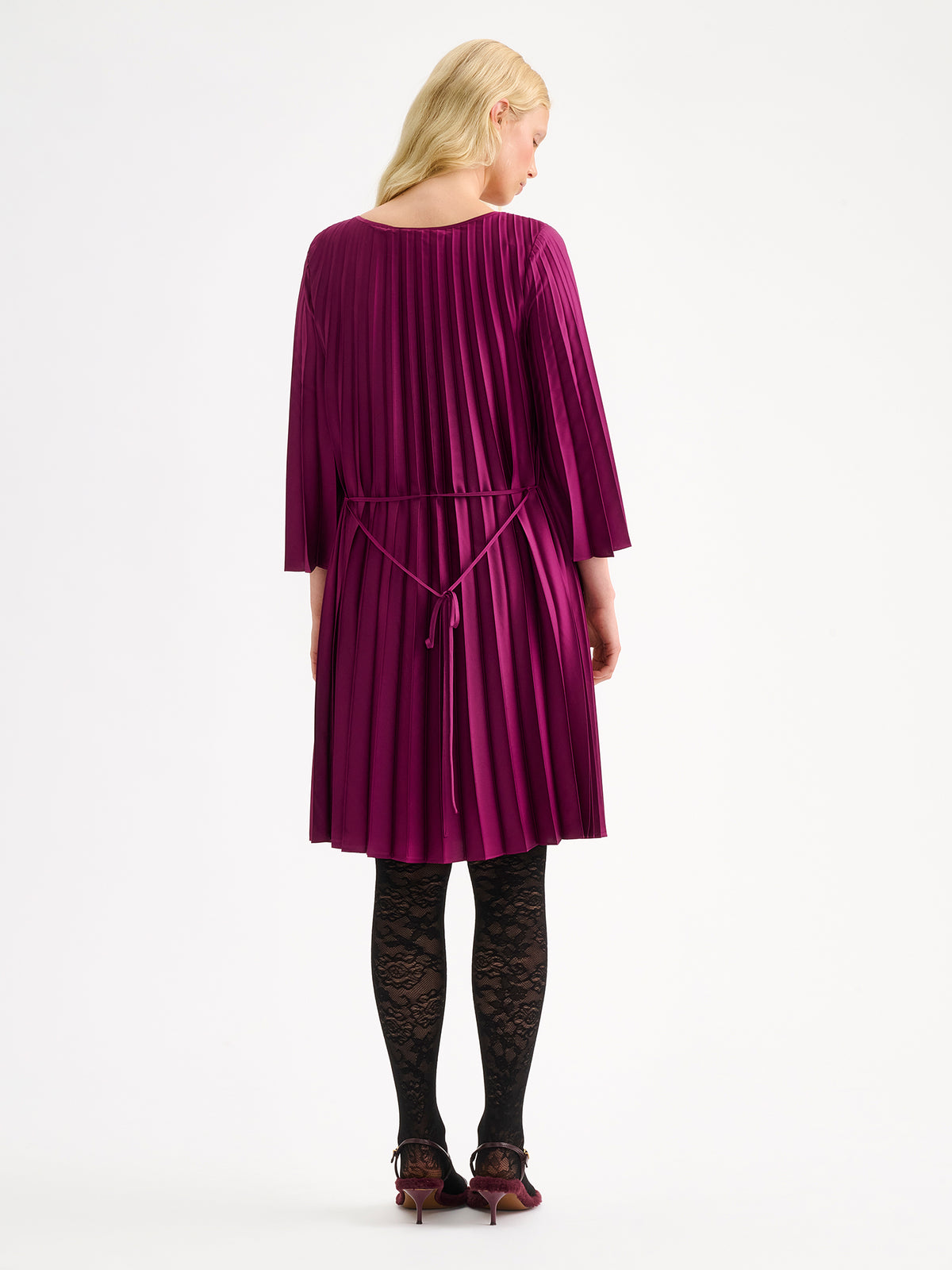Nanso women's beautifully draping Taite dress in a deliciously radiant violet shade. This versatile pleated dress can be worn with or without the slim belt. This A-line dress features 3/4-length sleeves and a slim belt at the waist. Pictured on a model with black lace tights from the backside.