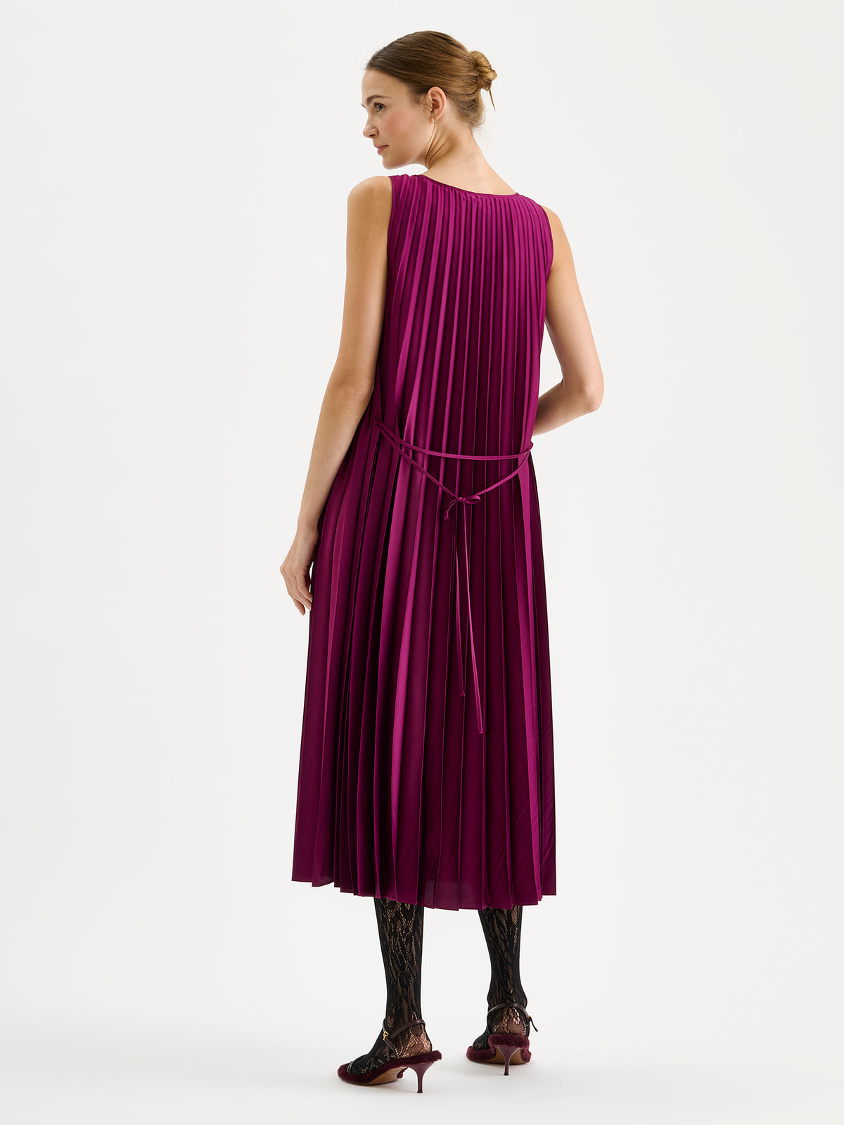 Nanso women's beautifully draping long Taite dress in a radiant violet shade. This long, sleeveless pleated dress with a slim belt is a striking choice for festive occasions and other special moments throughout the season. Pictured on a model with heels from the backside.