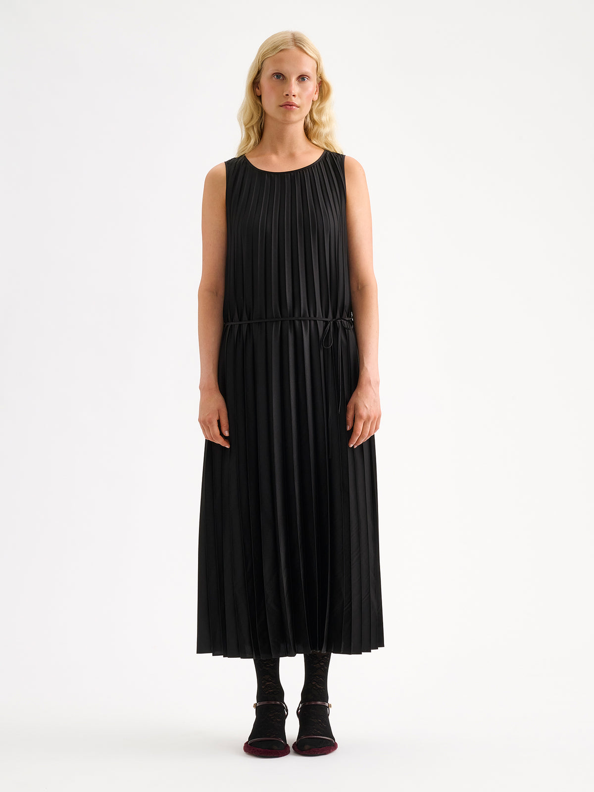 Nanso women's beautifully draping long Taite dress in a classic black shade. This long, sleeveless pleated dress with a slim belt is a striking choice for festive occasions and other special moments throughout the season. Pictured on a model close-up.