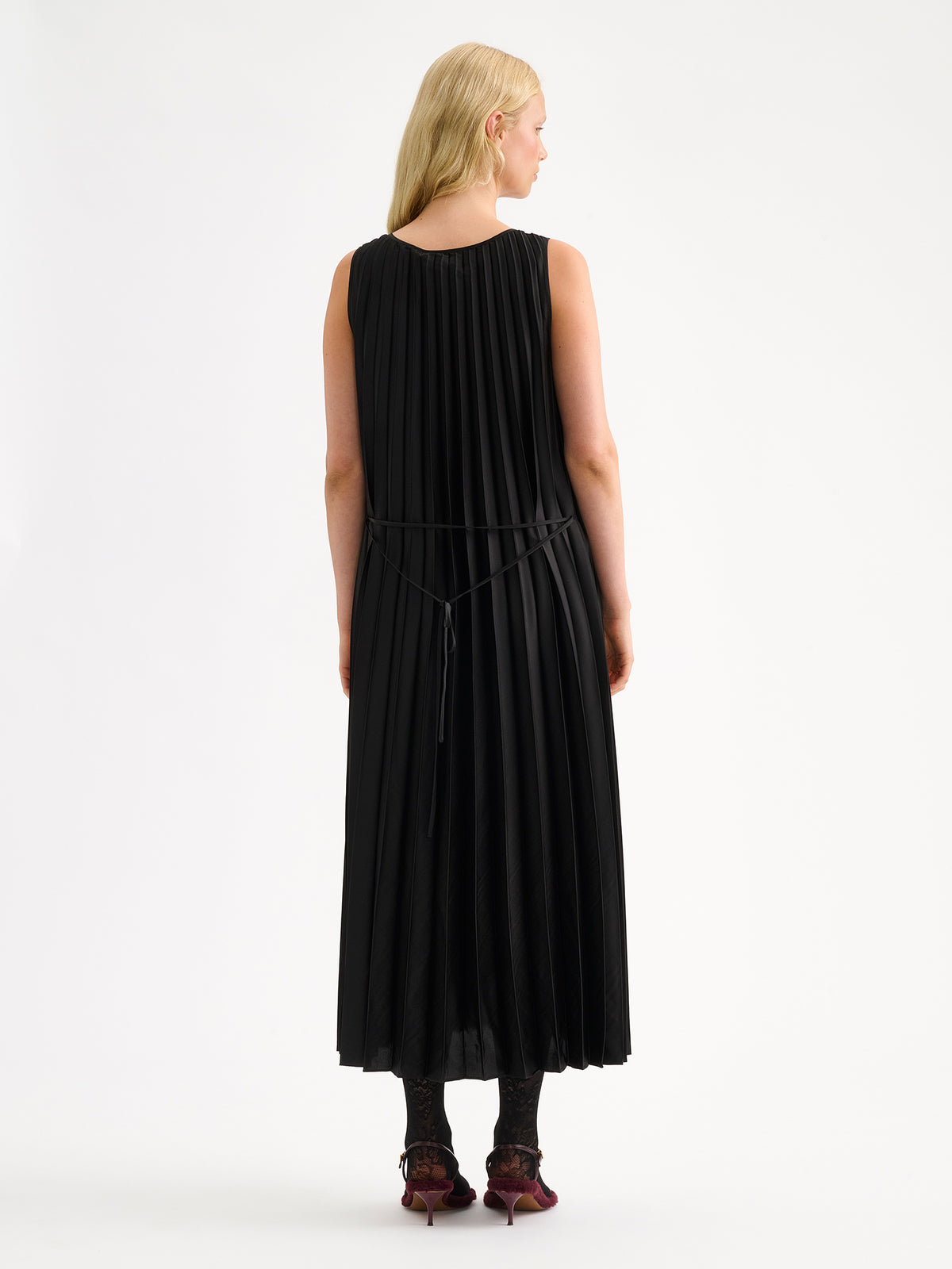 Nanso women's beautifully draping long Taite dress in a classic black shade. This long, sleeveless pleated dress with a slim belt is a striking choice for festive occasions and other special moments throughout the season. Pictured on a model from the backside.
