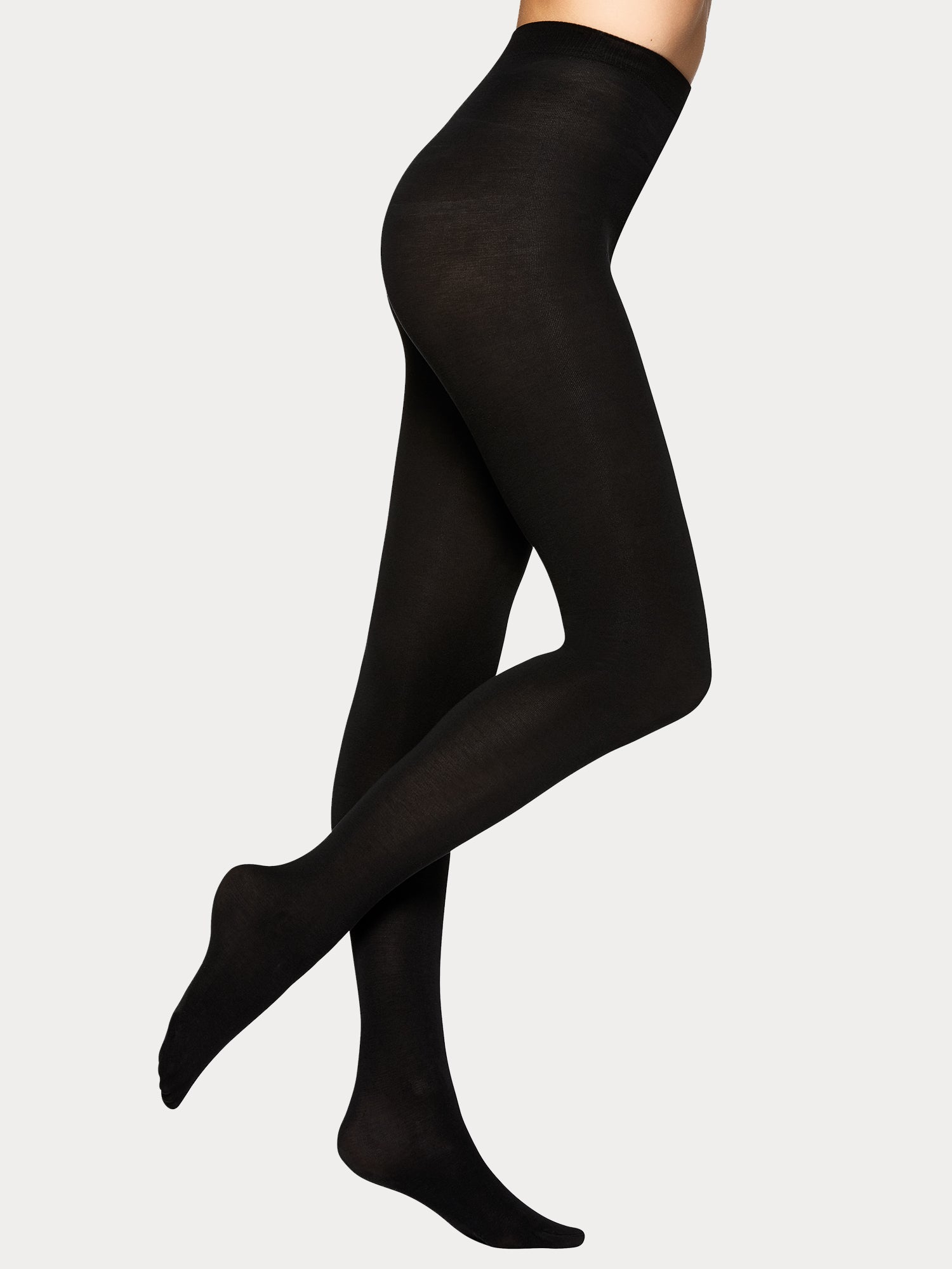 Vogue women's luxuriously soft black Silky Cotton tights crafted from organic cotton for everyday comfort with innovative 3D knit construction. On a model pictured from the side.
