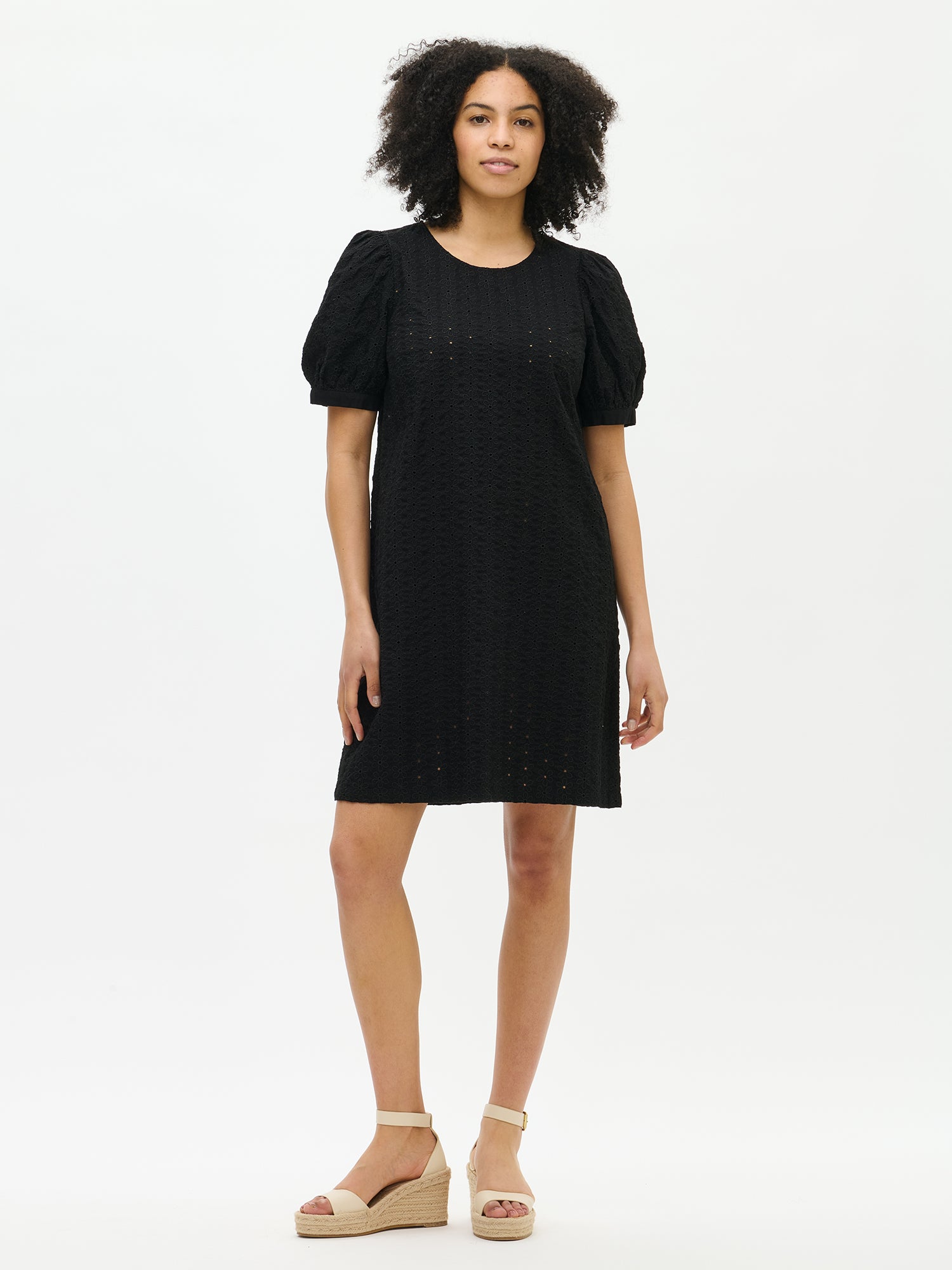 Nanso women's solid black Petra dress with a hem that reaches above the knees. The dress features a round neckline finished with a trim and bust darts. Shown in the image on a model from the front.