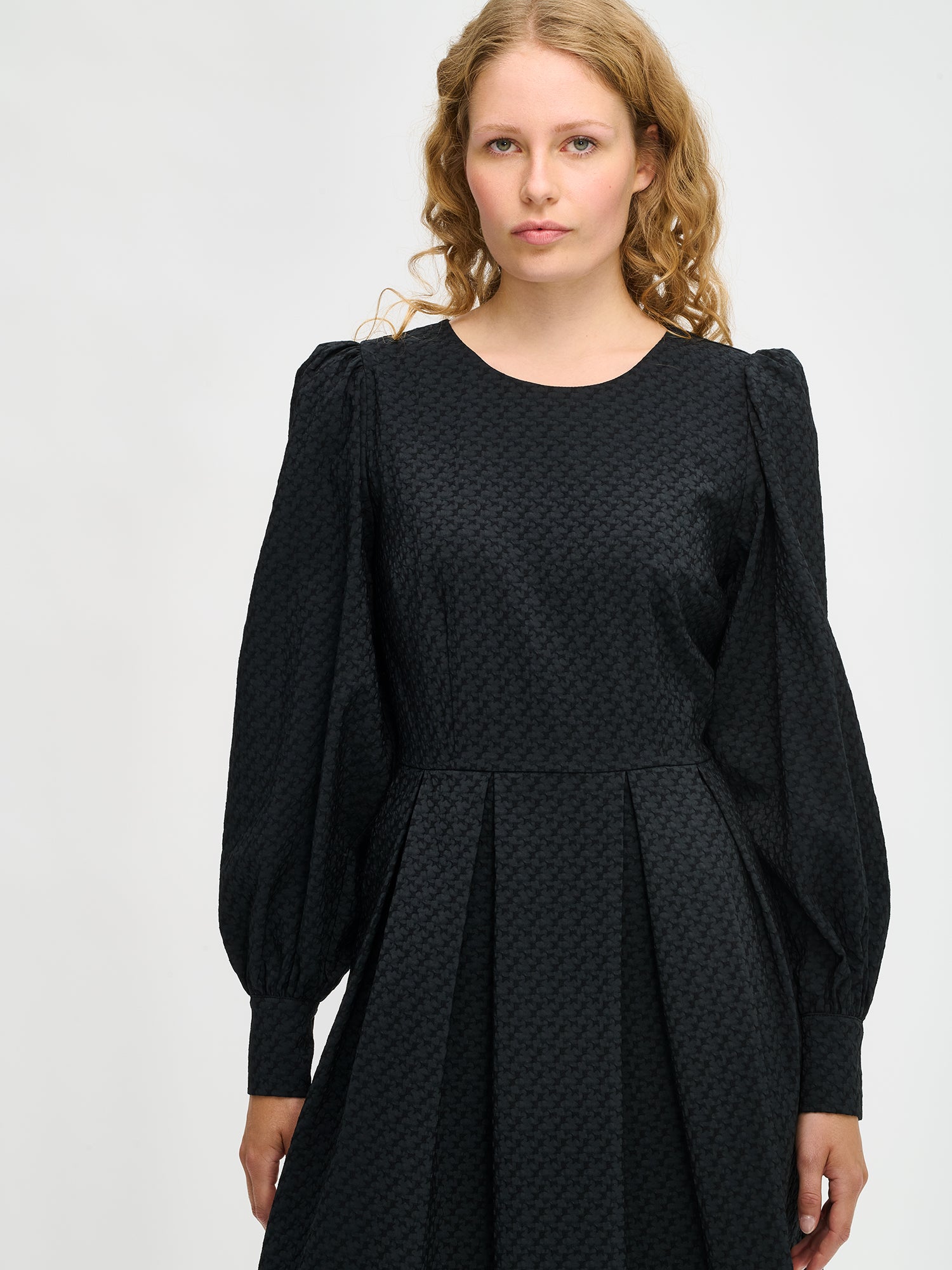 Nanso women’s black Reliefi dress, midi-length design with round neckline, gathered sleeves, pleated waist and side pockets, worn by model in close-up photo.
