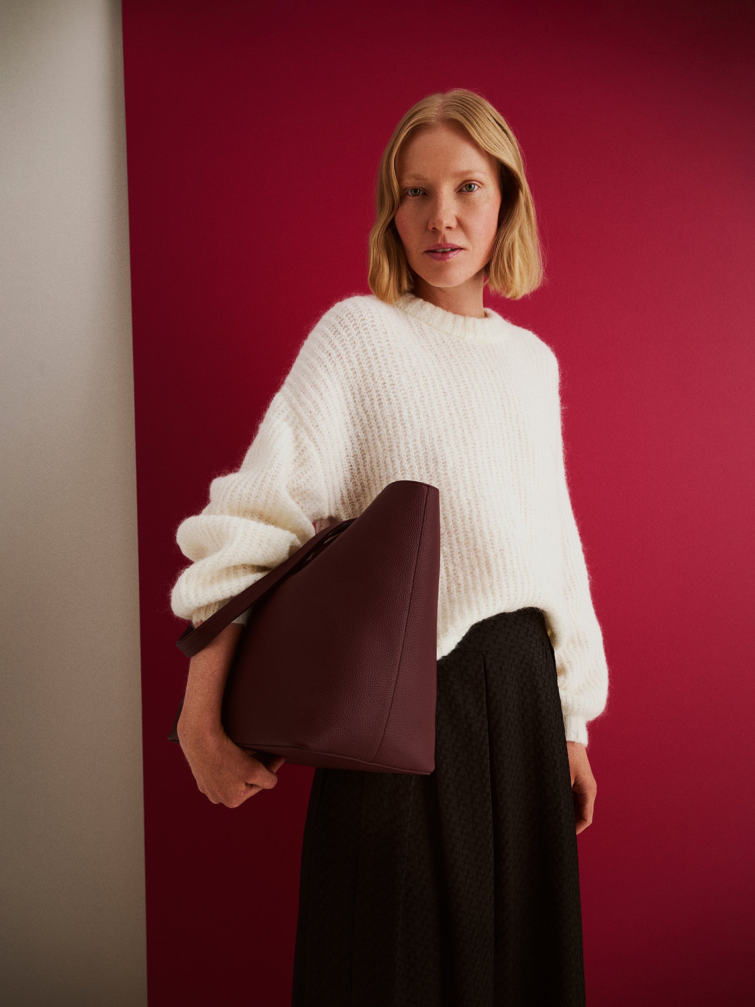 Nanso women’s black Reliefi skirt, midi-length style paired with white Karamelli knit sweater, worn by model in full-body photo against red background.
