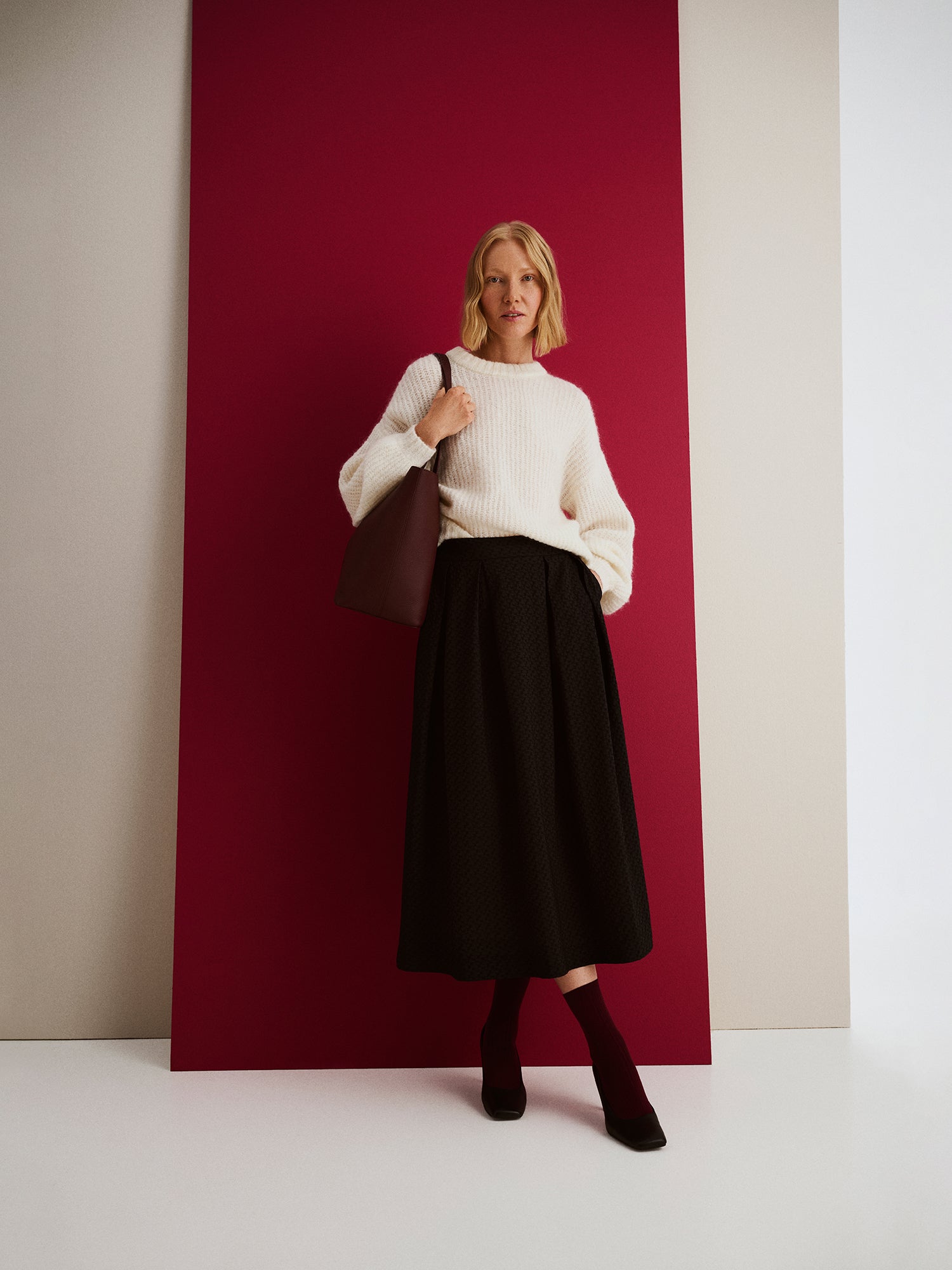 Nanso women’s black Reliefi skirt, midi-length style paired with white Karamelli knit sweater and brown Naru bag, worn by model in full-body photo.