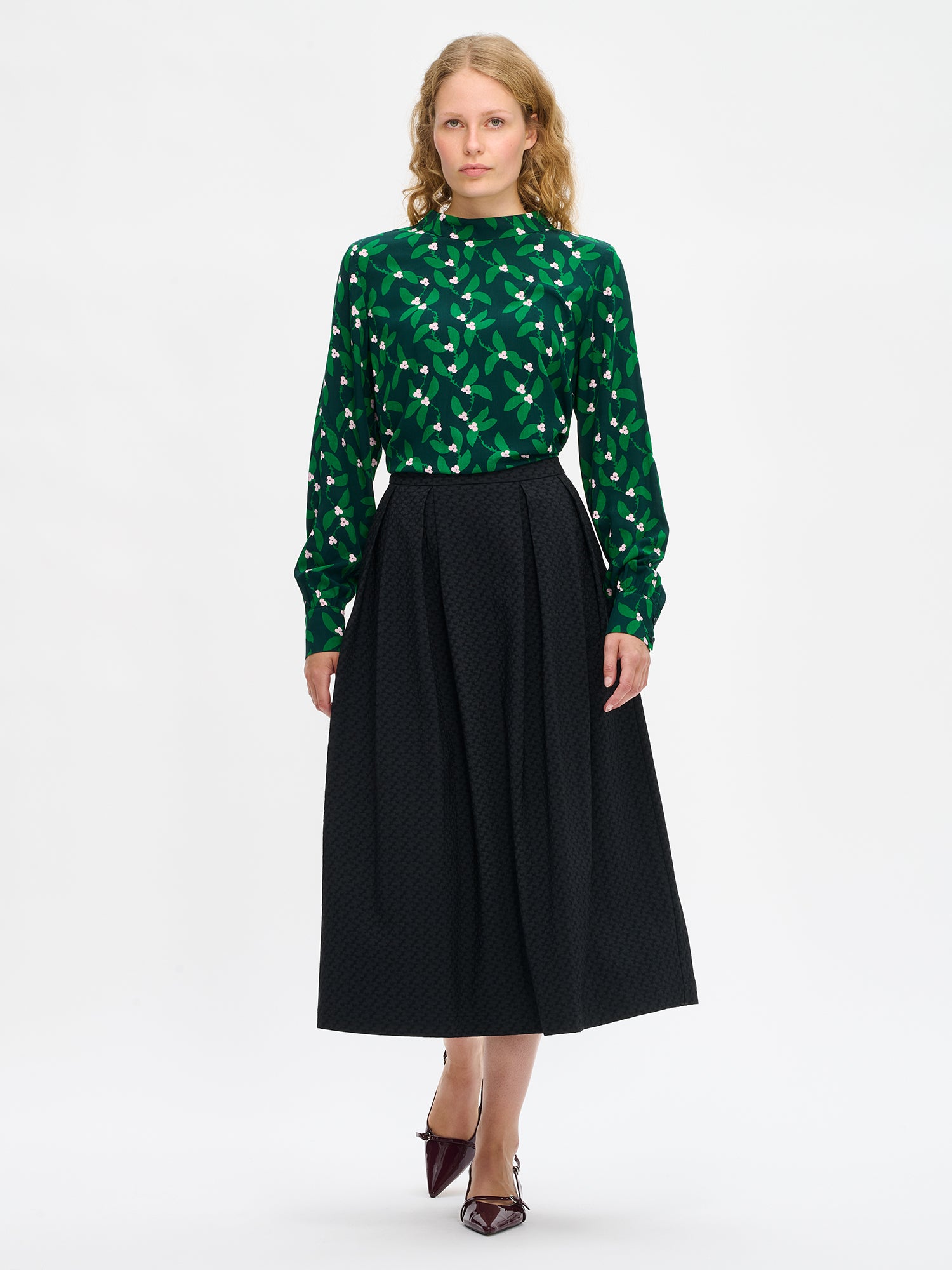 Nanso women’s black Reliefi skirt, midi-length style with pleats at the waist, gently flared hem and side pockets, worn by model with green top.
