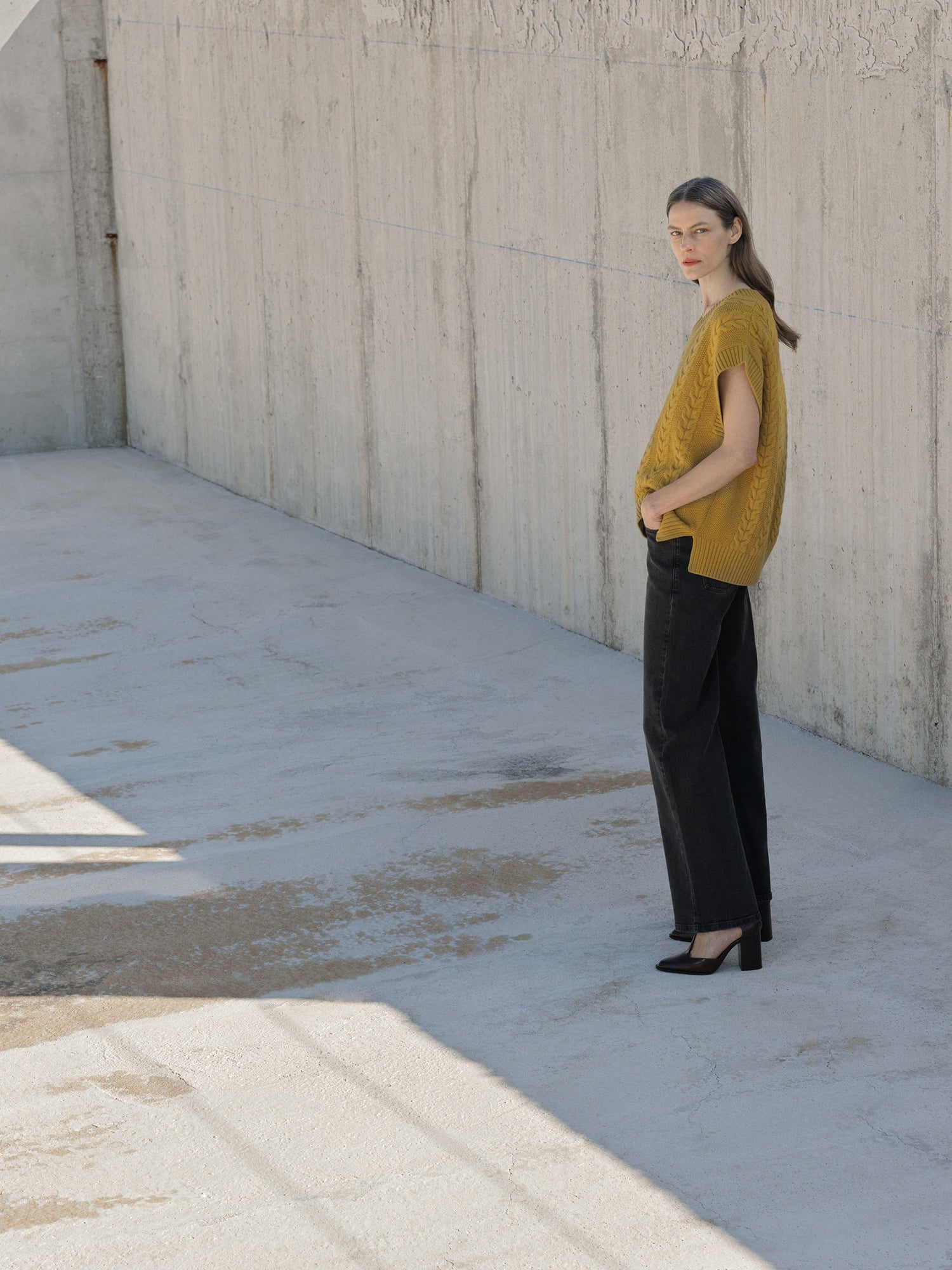 Woman in yellow knit vest and black pants standing against a concrete wall.