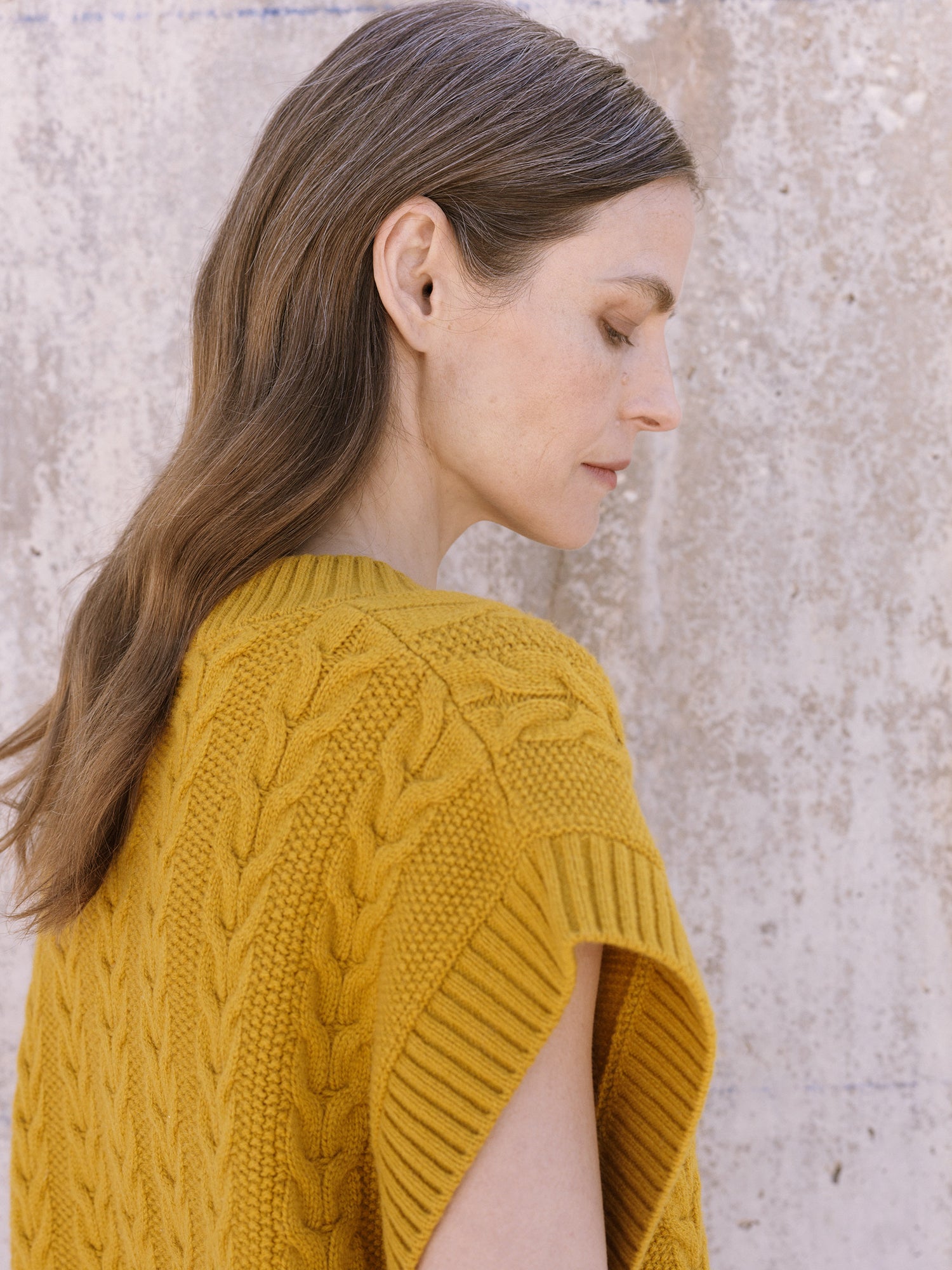 Woman wearing a yellow knitted vest against a stone wall.
