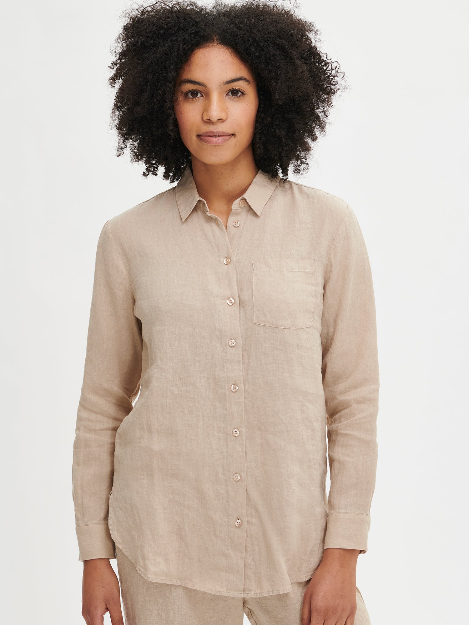 This beige PELLAVA linen shirt for women features full length sleeves, button-up front, and a front chest pocket. It is made of durable and breathable woven linen fabric. Worn by a model in size S from the front.