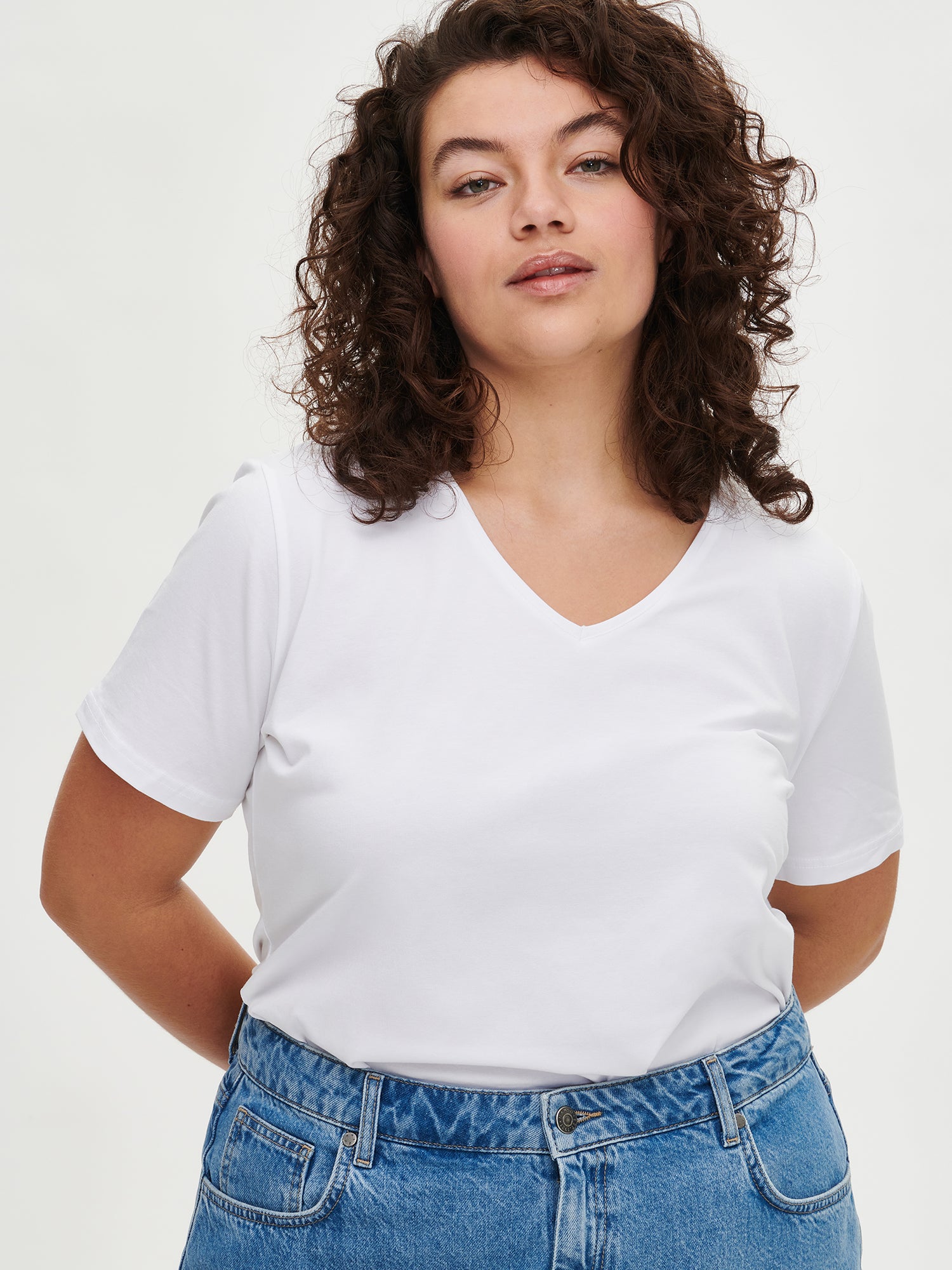 Women's Basic white T-shirt features a flattering V-neckline and comfortable short sleeves made from stretchy organic cotton-elastane blend, on a plus size model with classic blue jeans, close-up.
