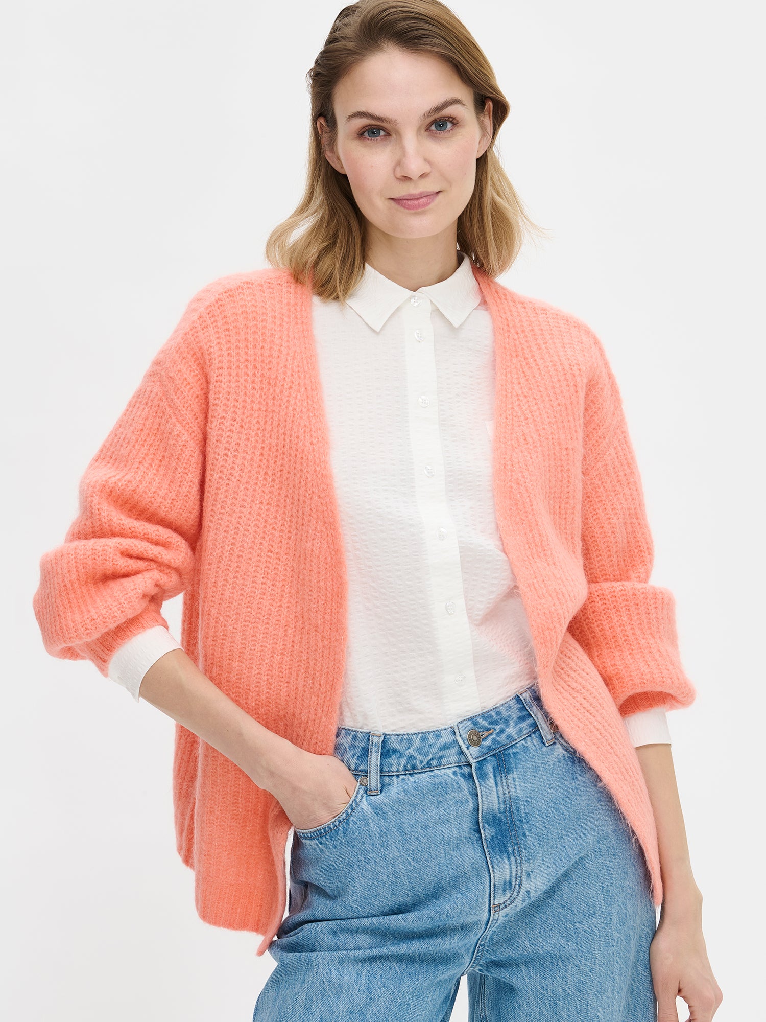 Women's long-sleeved KARAMELLI Cardigan in coral shade, knitted in a fitted shape. Made of a blend of certified mohair, certified wool, polyamide, and elastane. Pictured on a model with jeans, close-up.