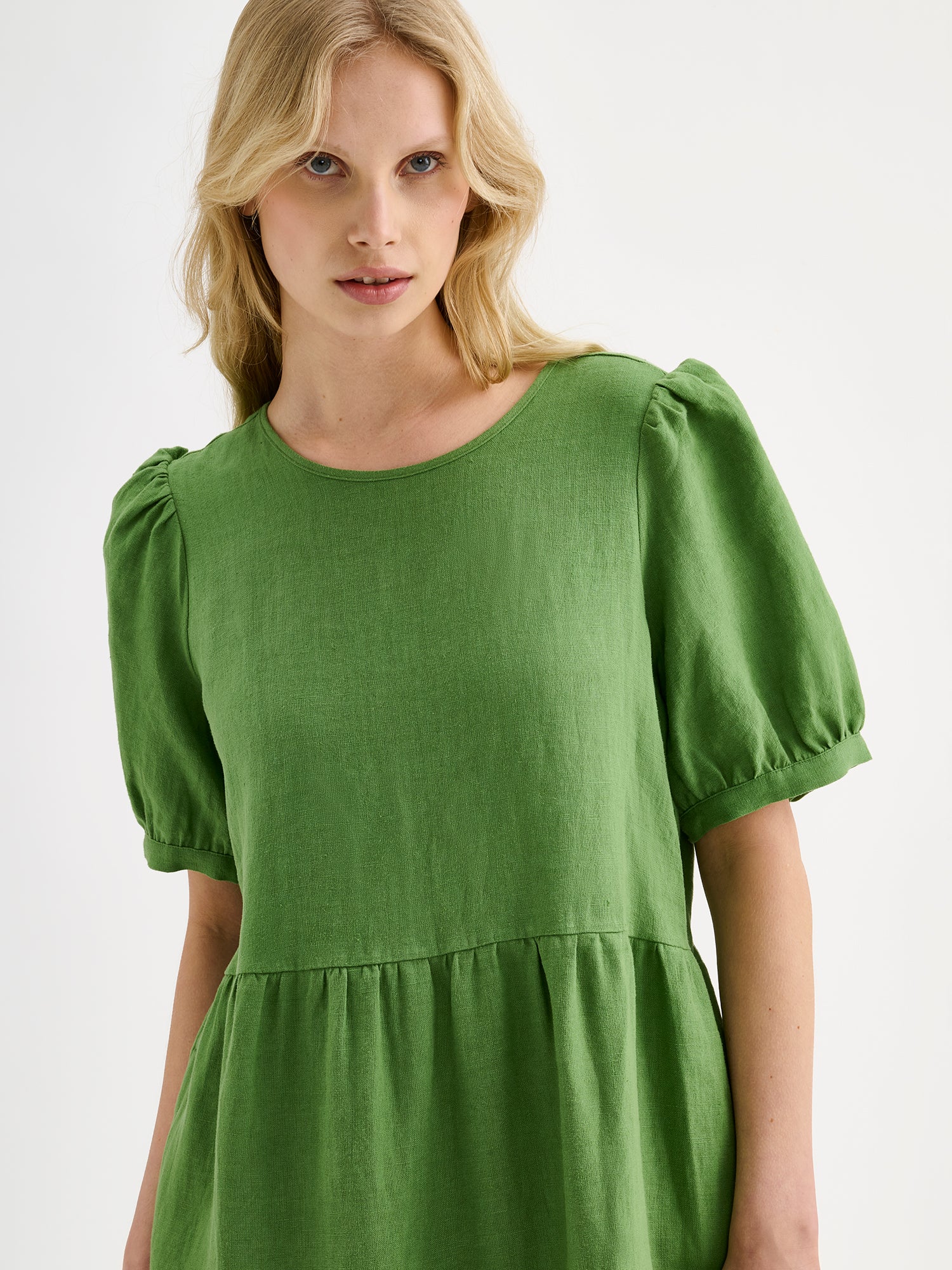 The Sarastus green dress for women is made of high-quality, durable, and breathable woven linen fabric. With its voluminous frill layers and short puff sleeves, this dress exudes both elegance and comfort. Pictured on a blonde model, close-up.