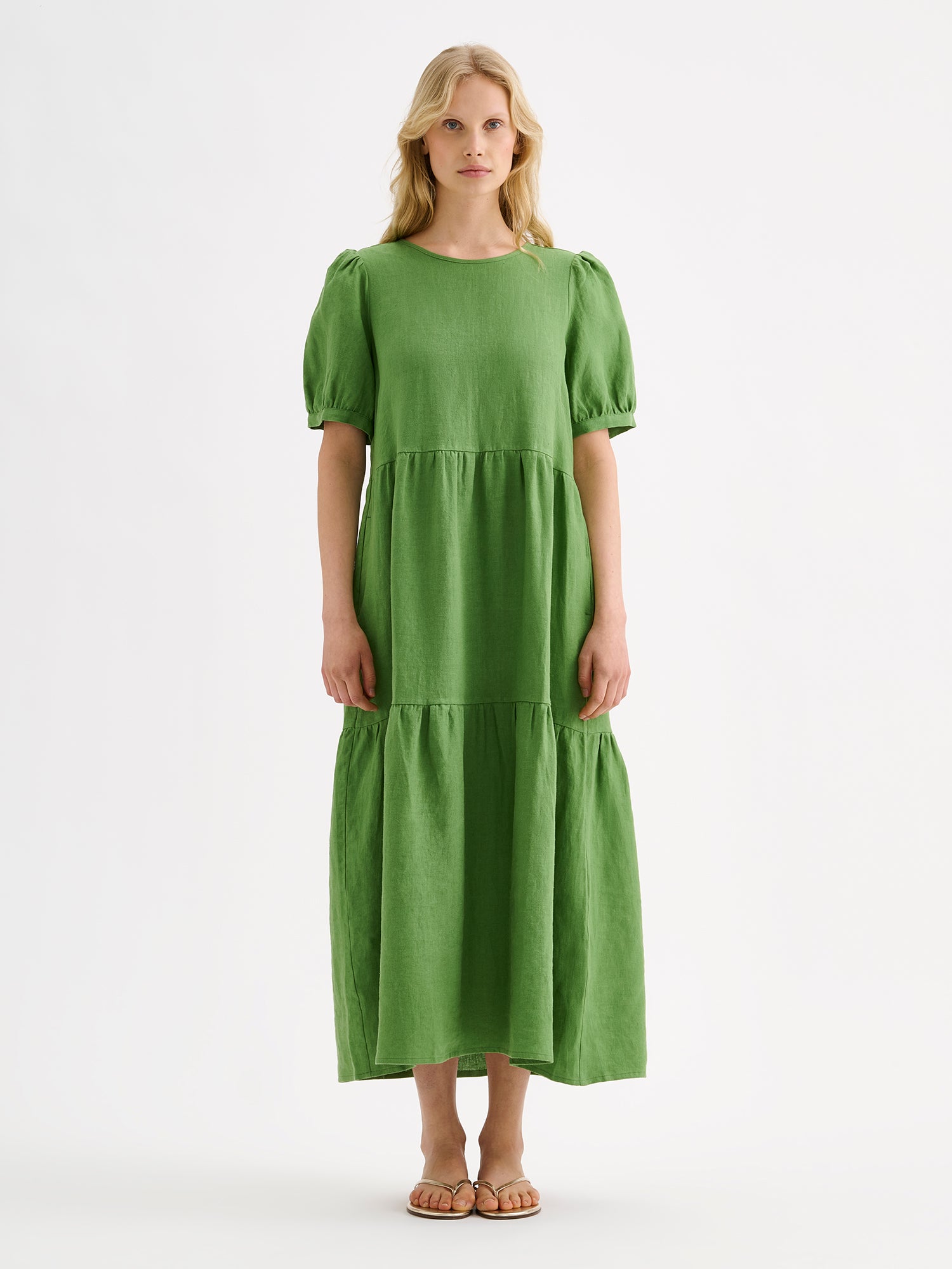 The Sarastus green dress for women is made of high-quality, durable, and breathable woven linen fabric. With its voluminous frill layers and short puff sleeves, this dress exudes both elegance and comfort. Pictured on a blonde model.