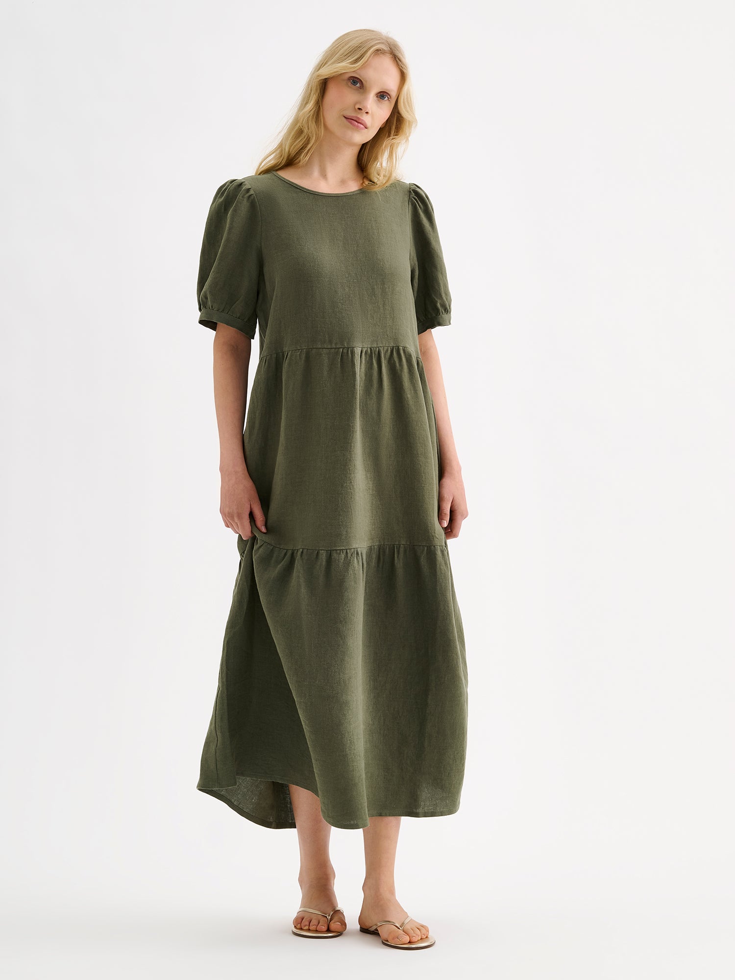 The Sarastus olive green dress for women is made of high-quality, durable, and breathable woven linen fabric. With its voluminous frill layers and short puff sleeves, this dress exudes both elegance and comfort. Pictured on a model with blonde hair.