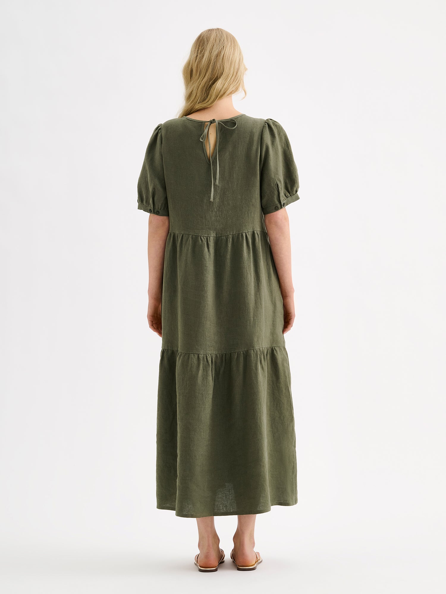 The Sarastus olive green dress for women is made of high-quality, durable, and breathable woven linen fabric. With its voluminous frill layers and short puff sleeves, this dress exudes both elegance and comfort. Pictured on a model with blonde hair, from the backside.