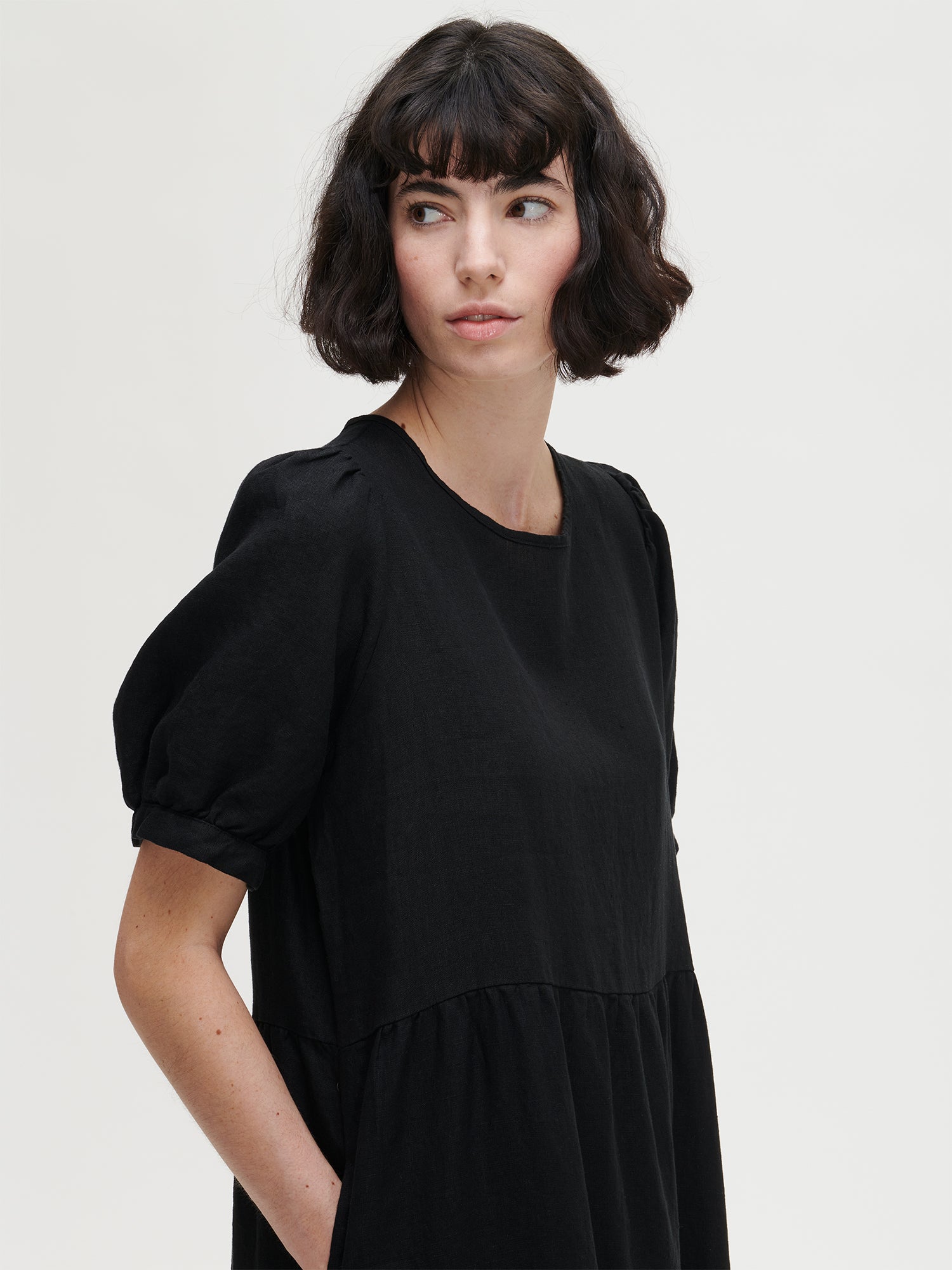 The Sarastus long black dress for women is made of high-quality, durable, and breathable woven linen fabric. With its voluminous frill layers and short puff sleeves, this dress exudes both elegance and comfort. Worn by a model in size S from the side.