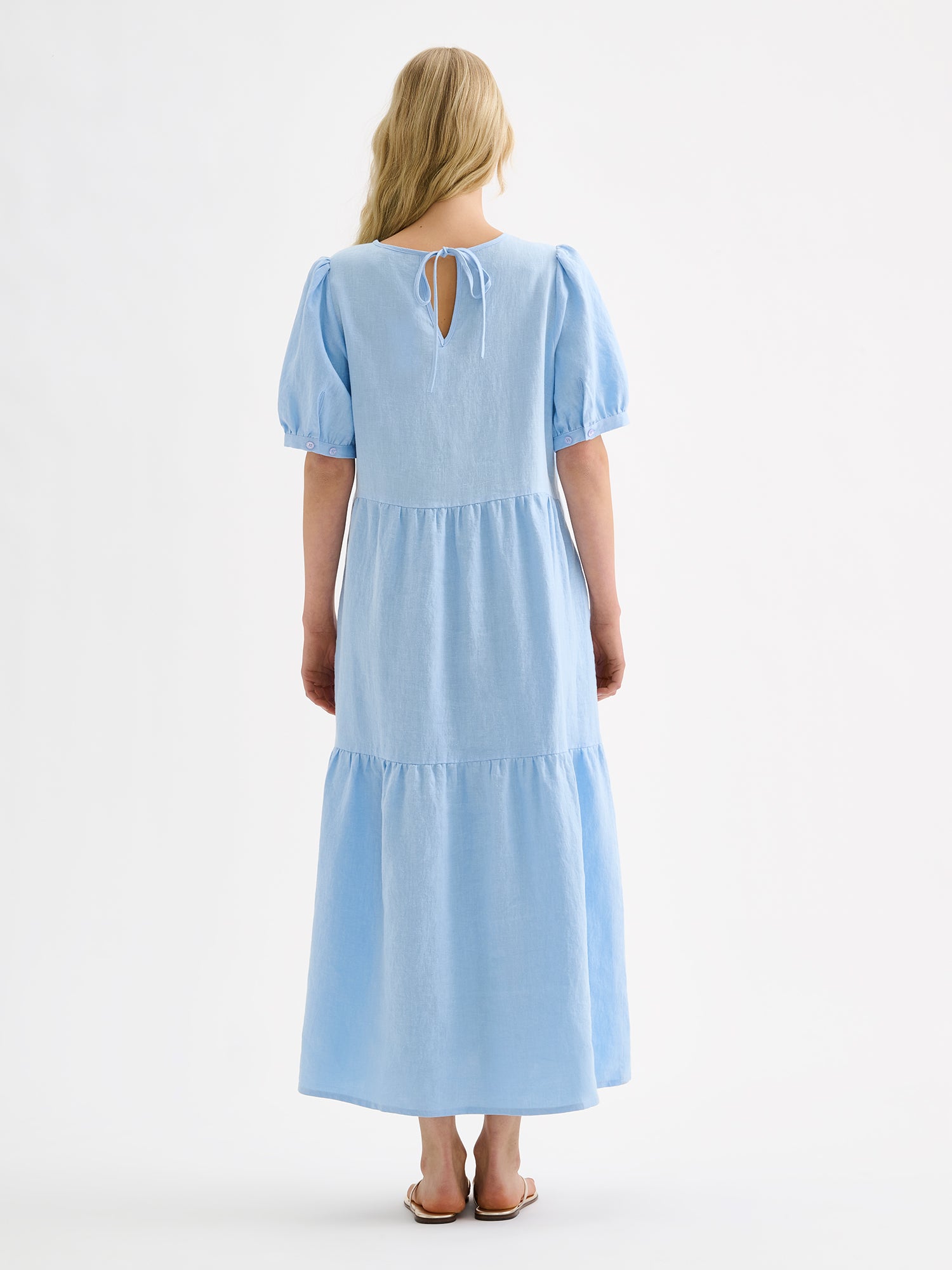 The Sarastus long soft light blue dress for women is made of high-quality, durable, and breathable woven linen fabric. With its voluminous frill layers and short puff sleeves, this dress exudes both elegance and comfort. Pictured on a blonde model, from the backside