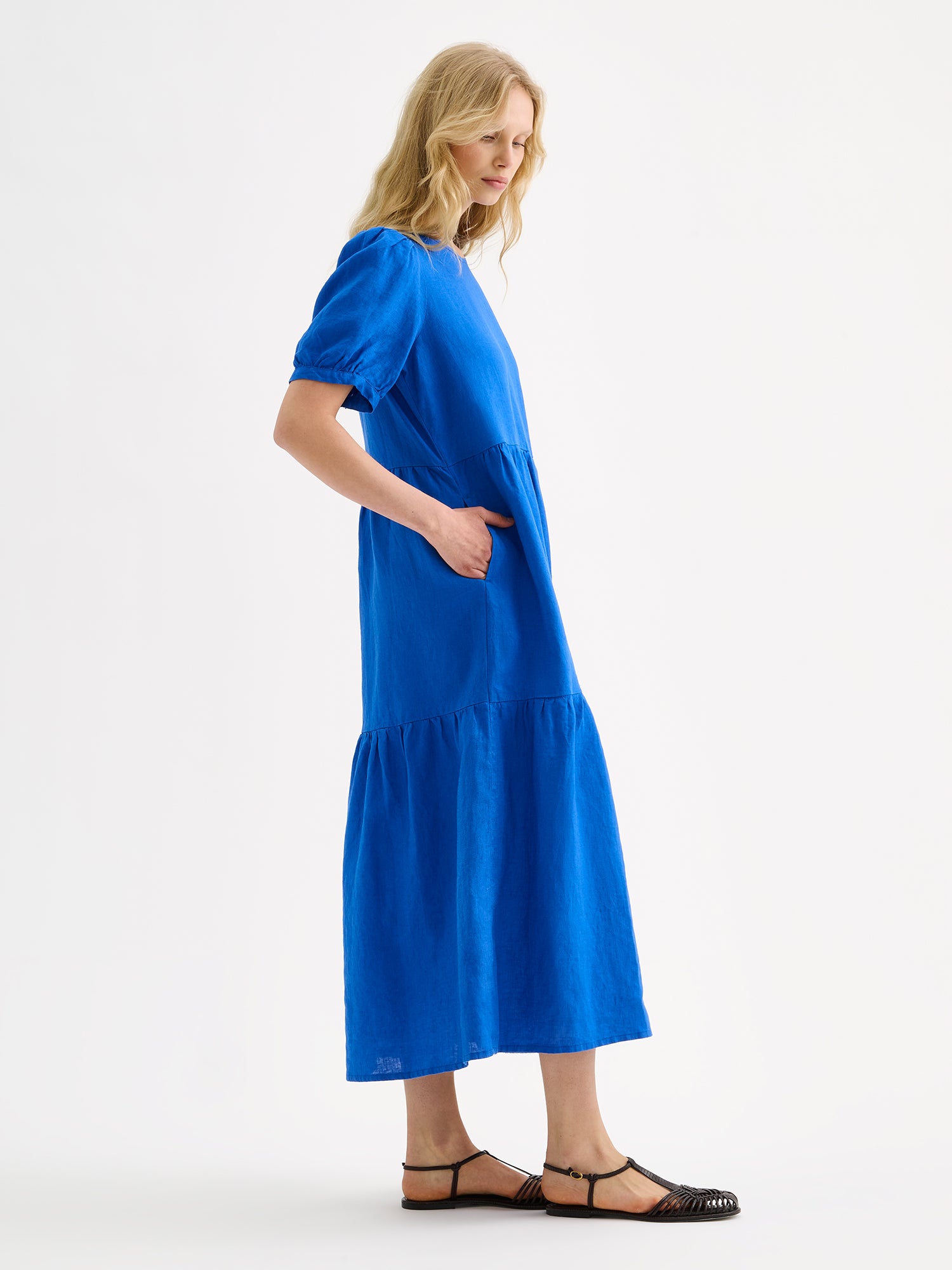 The Sarastus long blue dress for women is made of high-quality, durable, and breathable woven linen fabric, on a model with sandals, pictured from the side angle.