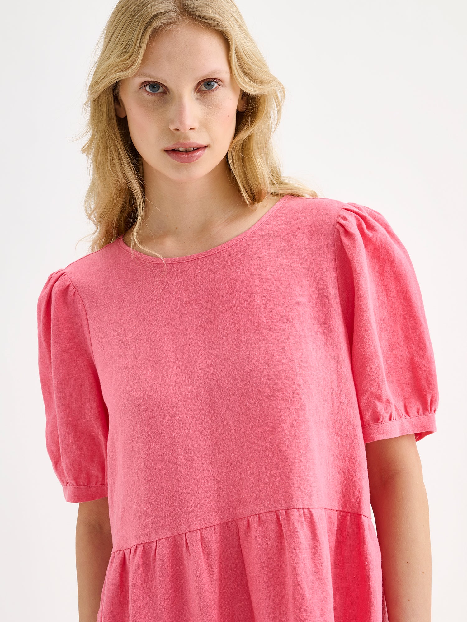 The Sarastus pink dress for women is made of high-quality, durable, and breathable woven linen fabric. With its voluminous frill layers and short puff sleeves, this dress exudes both elegance and comfort. Pictured on a blonde model, close-up.