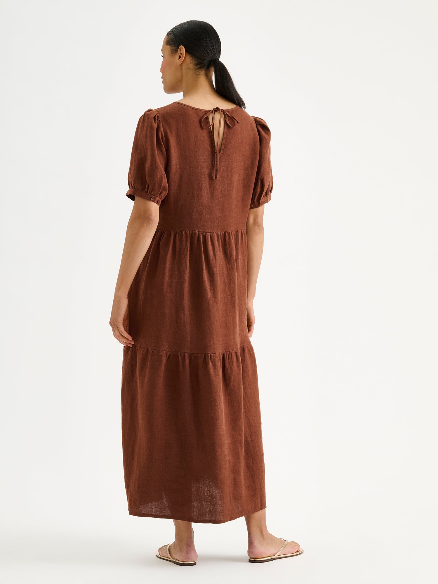 The Sarastus long brown linen dress with voluminous frill layers and short puff sleeves on a model, pictured from the backside.