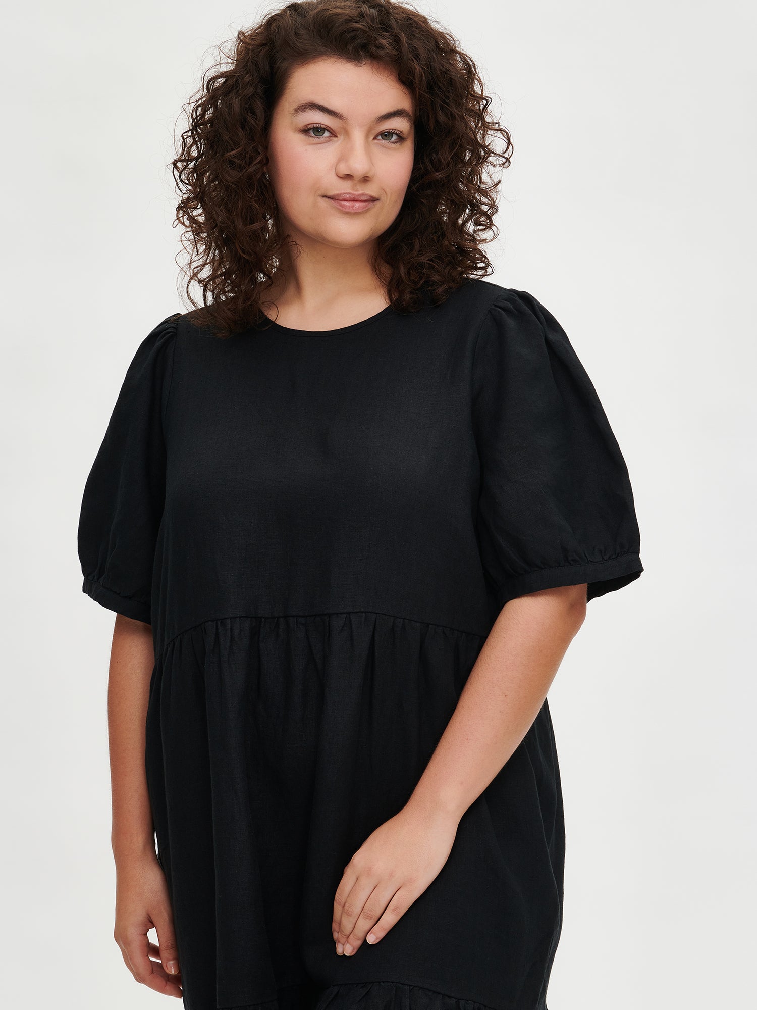 The Sarastus long black dress for women is made of high-quality, durable, and breathable woven linen fabric. With its voluminous frill layers and short puff sleeves, this dress exudes both elegance and comfort. Worn by a plus-size model in size XL from the front.