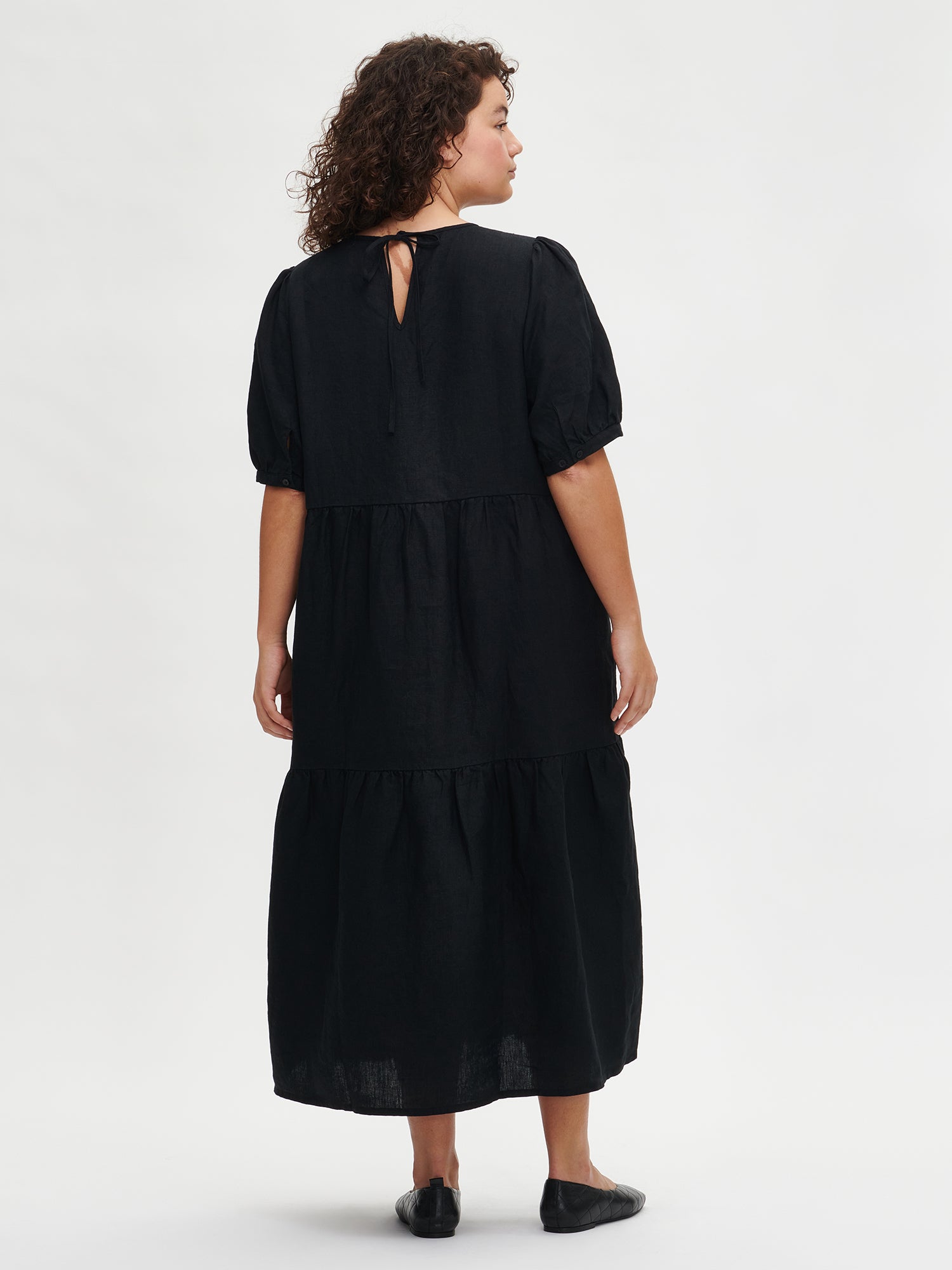 The Sarastus long black dress for women is made of high-quality, durable, and breathable woven linen fabric. With its voluminous frill layers and short puff sleeves, this dress exudes both elegance and comfort. Worn by a plus-size model in size XL from the back.