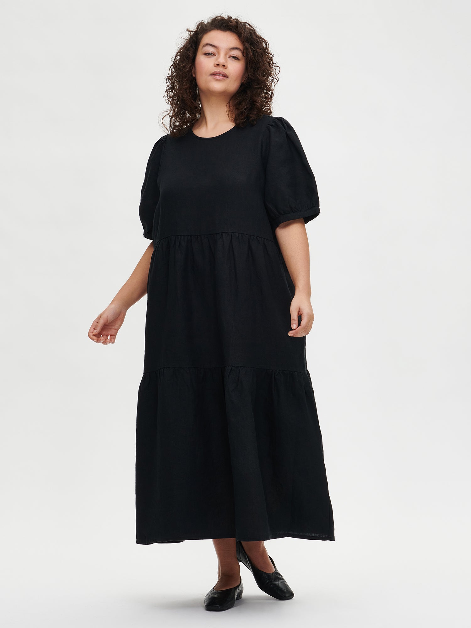 The Sarastus long black dress for women is made of high-quality, durable, and breathable woven linen fabric. With its voluminous frill layers and short puff sleeves, this dress exudes both elegance and comfort. Worn by a plus-size model in size XL from the front.