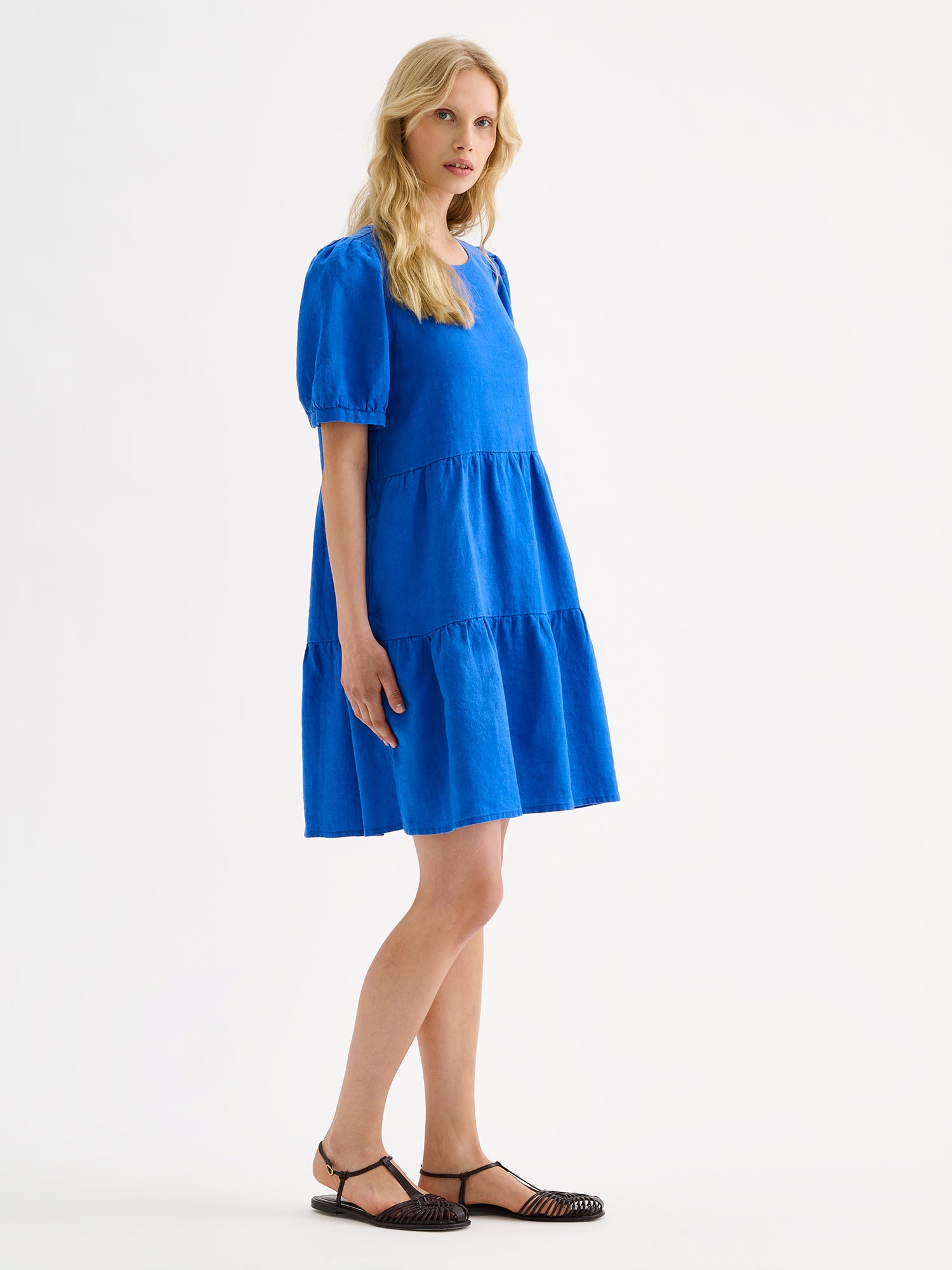 Nanso women's beautiful blue Sarastus linen dress with voluminous frill layers and short puff sleeves, on a model with sandals, pictured from the side angle.