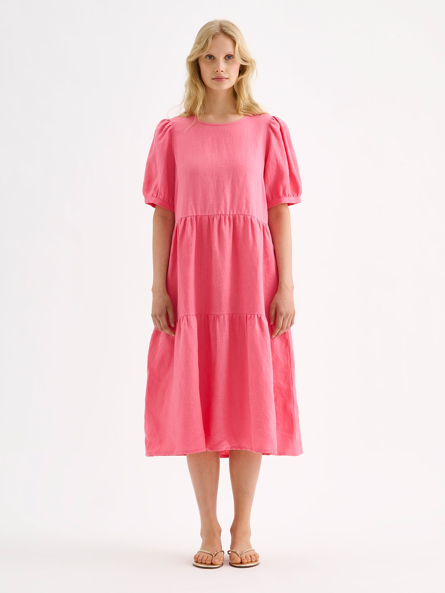 Nanso women's beautiful pink Sarastus midi linen dress features voluminous frill layers and short puff sleeves. This stylish summer dress is made of high-quality linen. Pictured on a blonde model with sandals.
