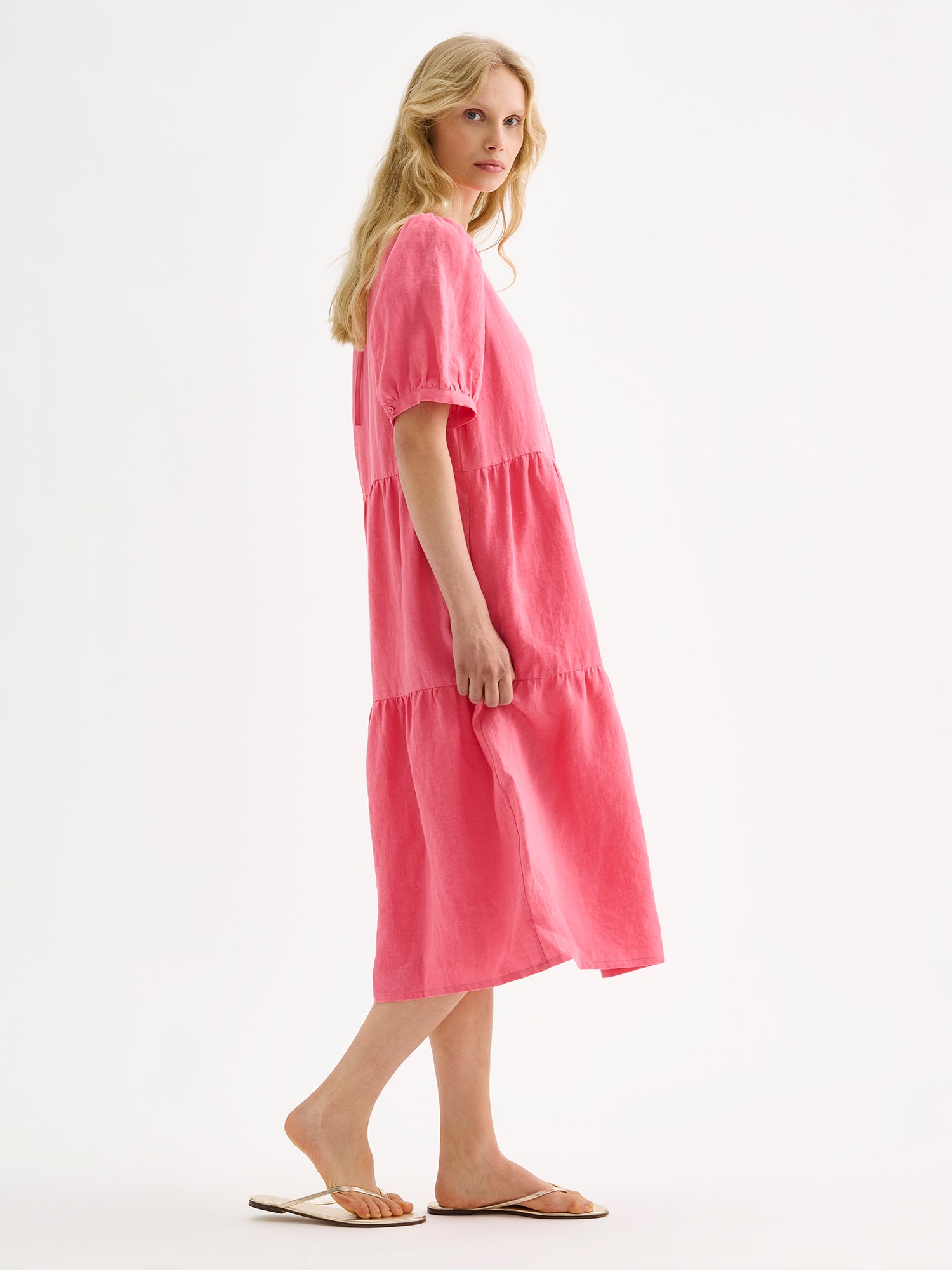 Nanso women's beautiful pink Sarastus midi linen dress features voluminous frill layers and short puff sleeves. This stylish summer dress is made of high-quality linen. Pictured from a side angle  on a blonde model with sandals.