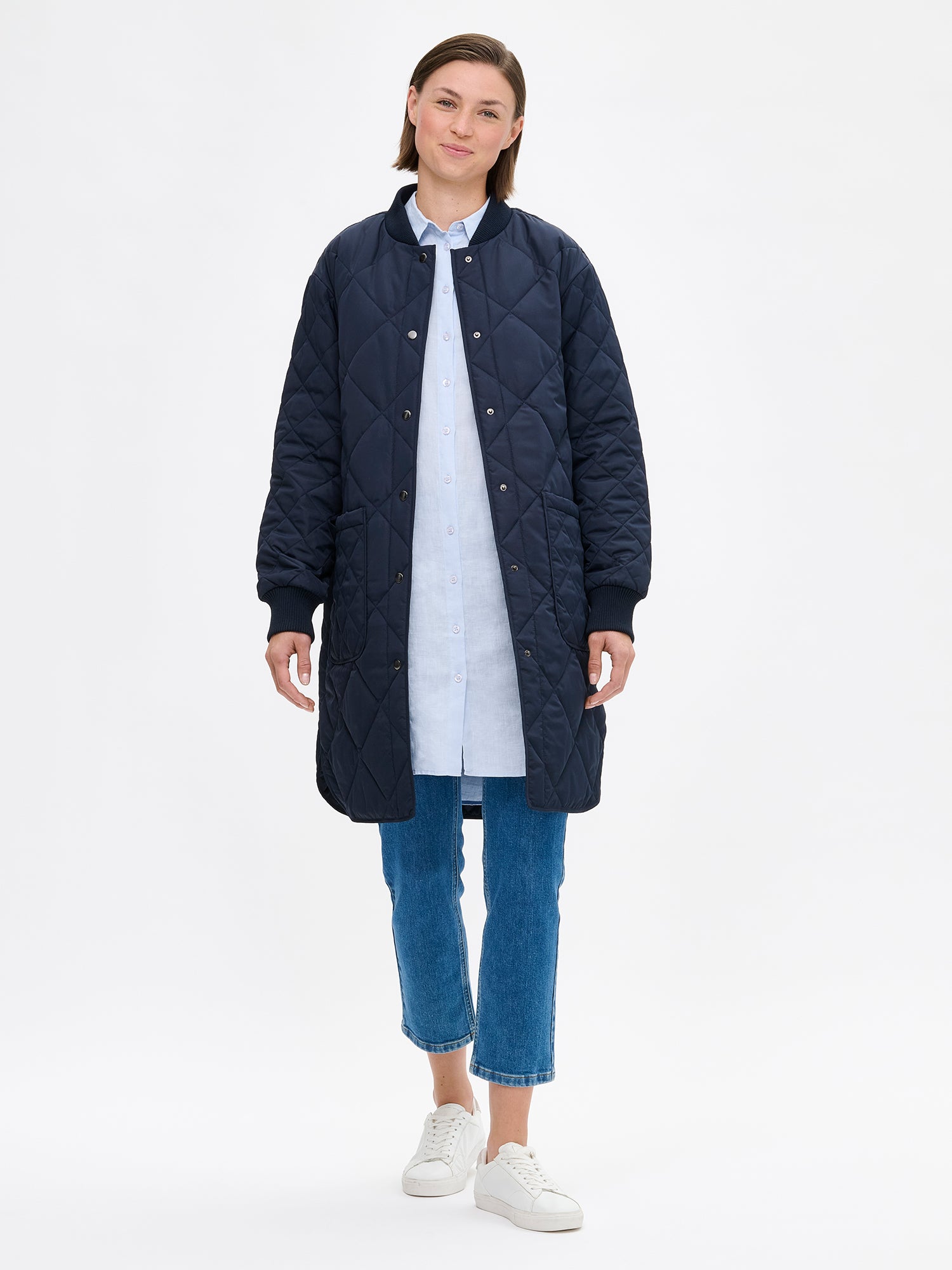 Nanso women's casual Midi quilted coat in dark blue features lightweight padding, ribbed knit detailing at the neckline and cuffs, snap closures and front pockets. Pictured on a model with jeans and sneakers, the coat opened.