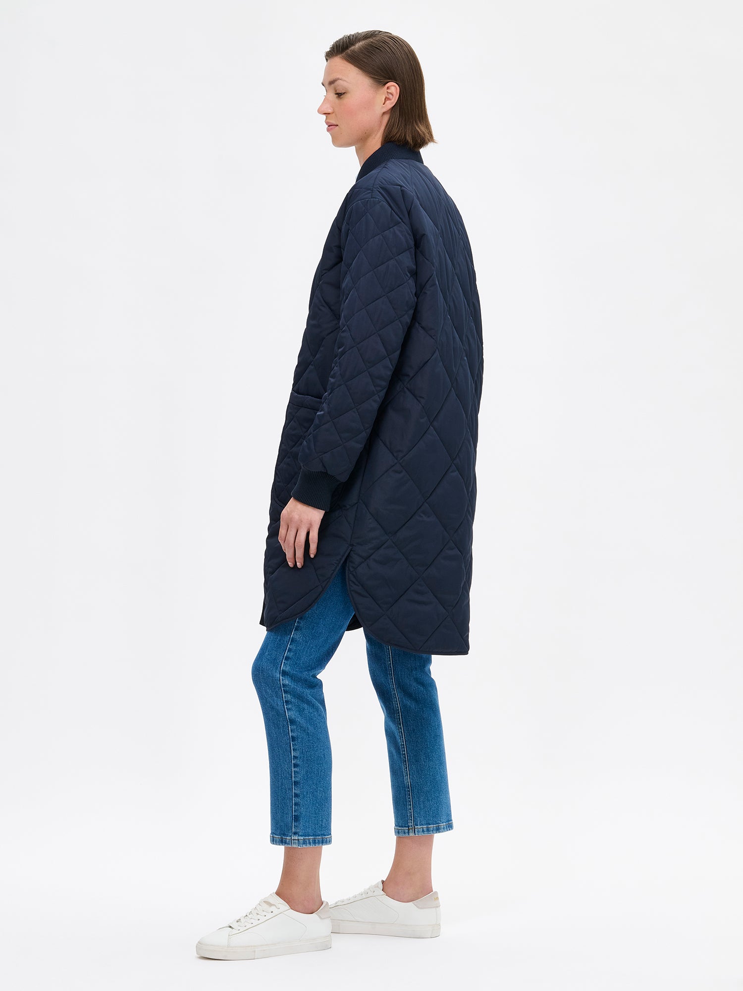 Nanso women's casual Midi quilted coat in dark blue features lightweight padding, ribbed knit detailing at the neckline and cuffs, snap closures and front pockets. Pictured on a model with jeans and sneakers, from the side view.