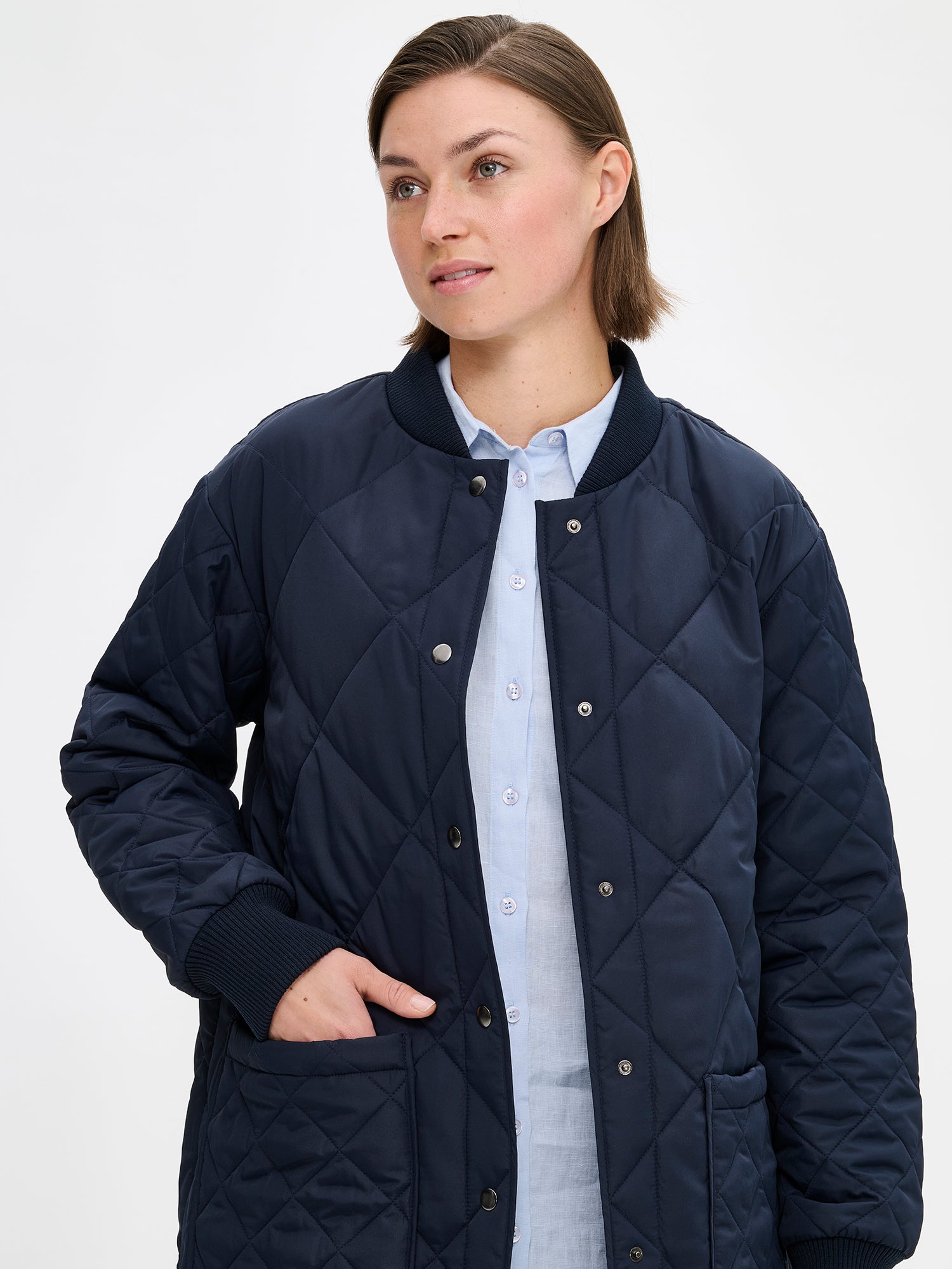 Nanso women's casual Midi quilted coat in dark blue features lightweight padding, ribbed knit detailing at the neckline and cuffs, snap closures and front pockets. Pictured on a model with jeans and sneakers, the coat opened, close-up.