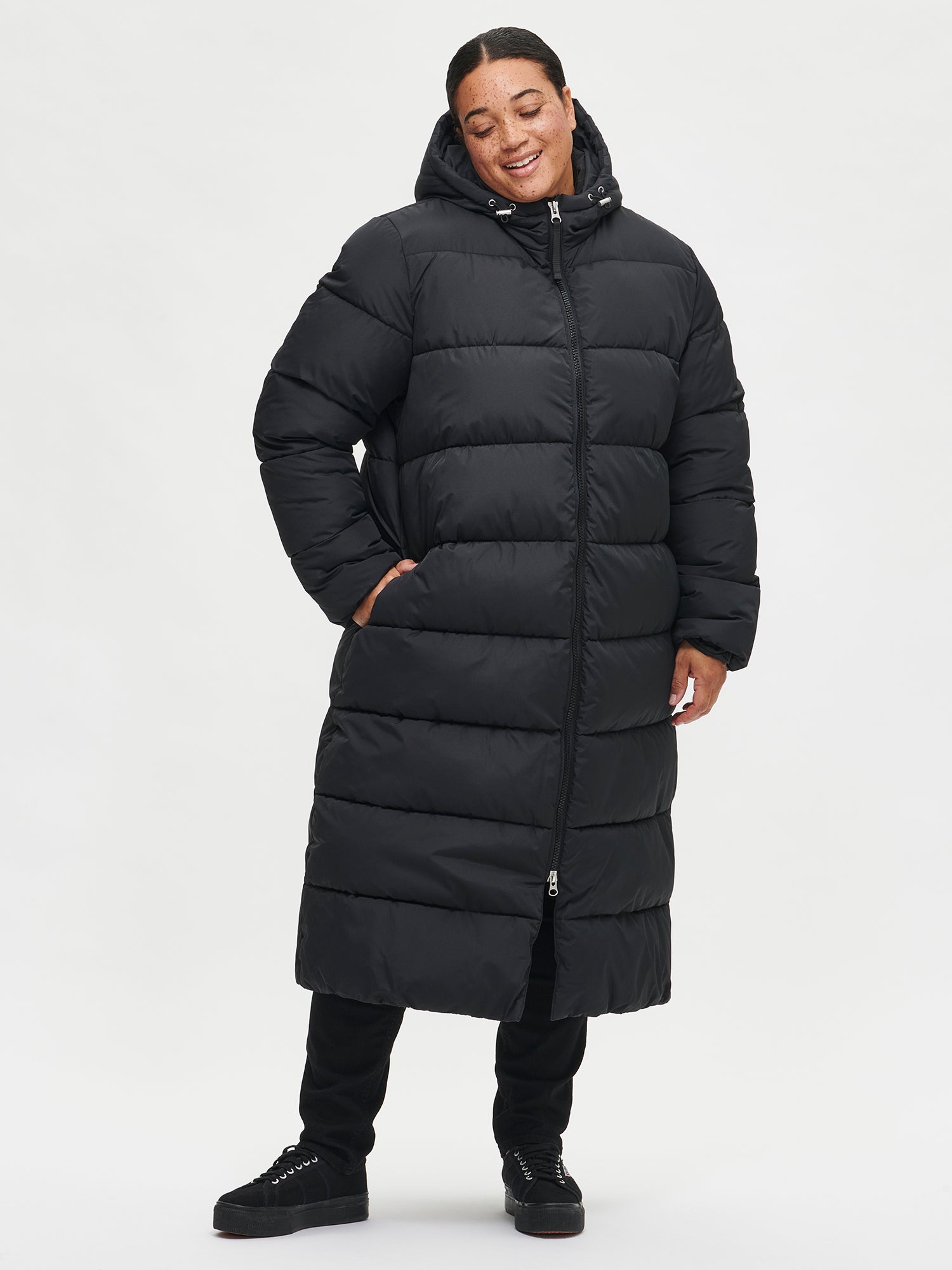 The Lumi Long Pink Puffer Coat in black offers cozy warmth with its lightweight, puffy insulation. Worn by a model in size S.