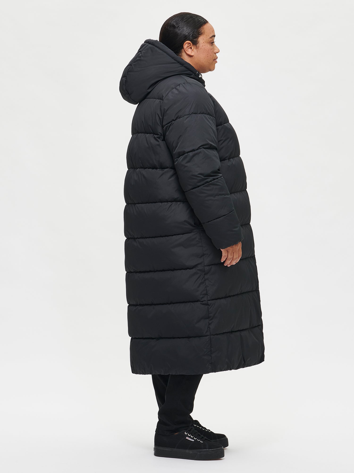 The Lumi Long Pink Puffer Coat in black offers cozy warmth with its lightweight, puffy insulation. Worn by a model from the side.