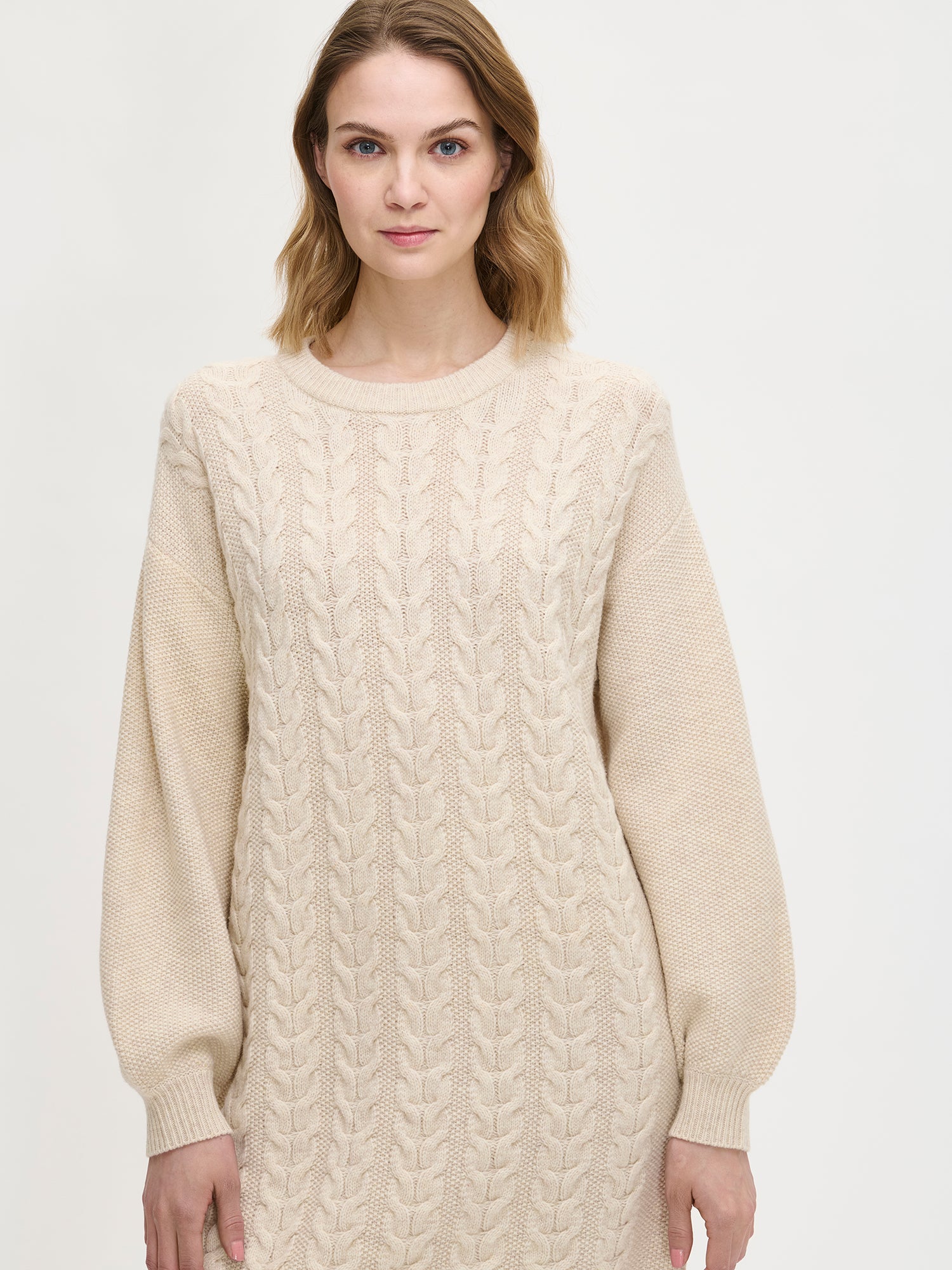 Woman wearing a beige cable knit sweater dress against a white background