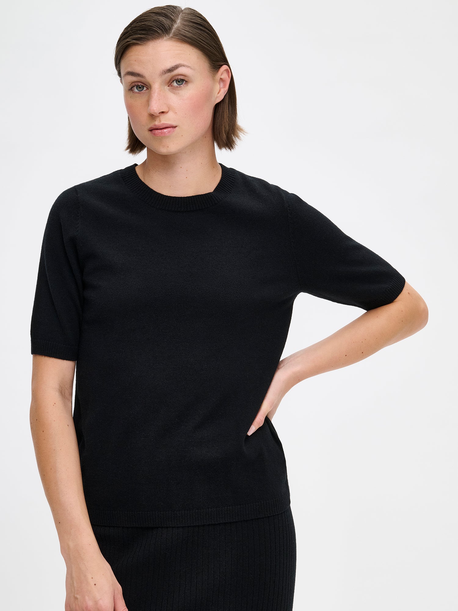 Nanso Villis women's short-sleeved black knit sweater features a classic crew neckline with half-length sleeves finished with ribbed trim at the cuffs. On a model with Villis ribbed skirt, close-up.