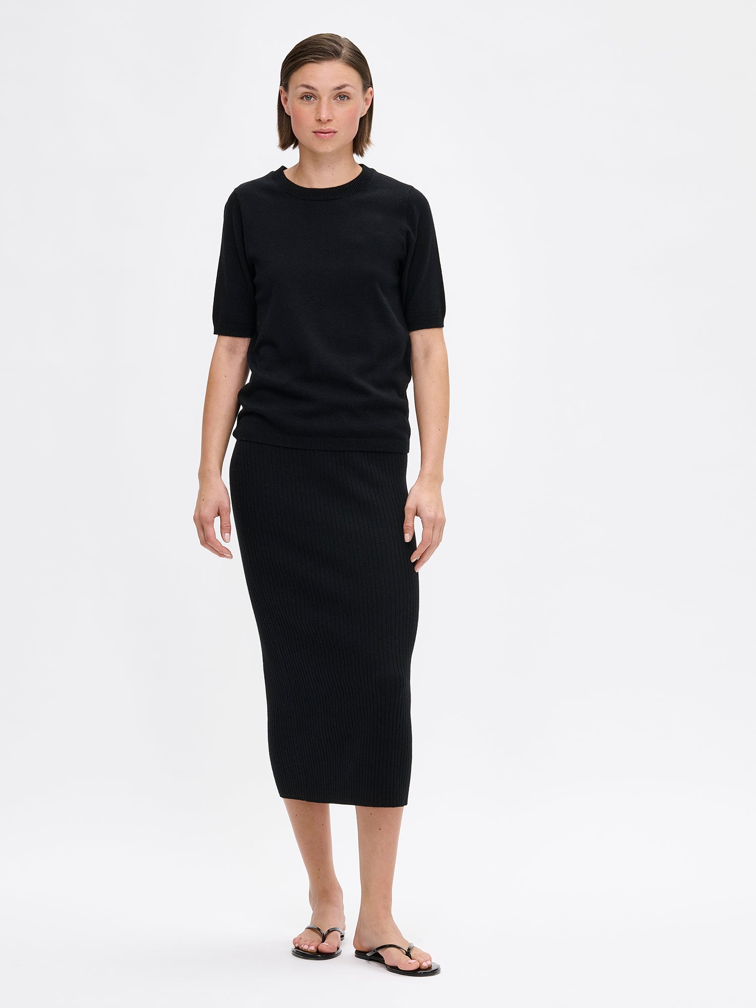 Nanso Villis women's short-sleeved black knit sweater features a classic crew neckline with half-length sleeves finished with ribbed trim at the cuffs. On a model with Villis ribbed skirt.