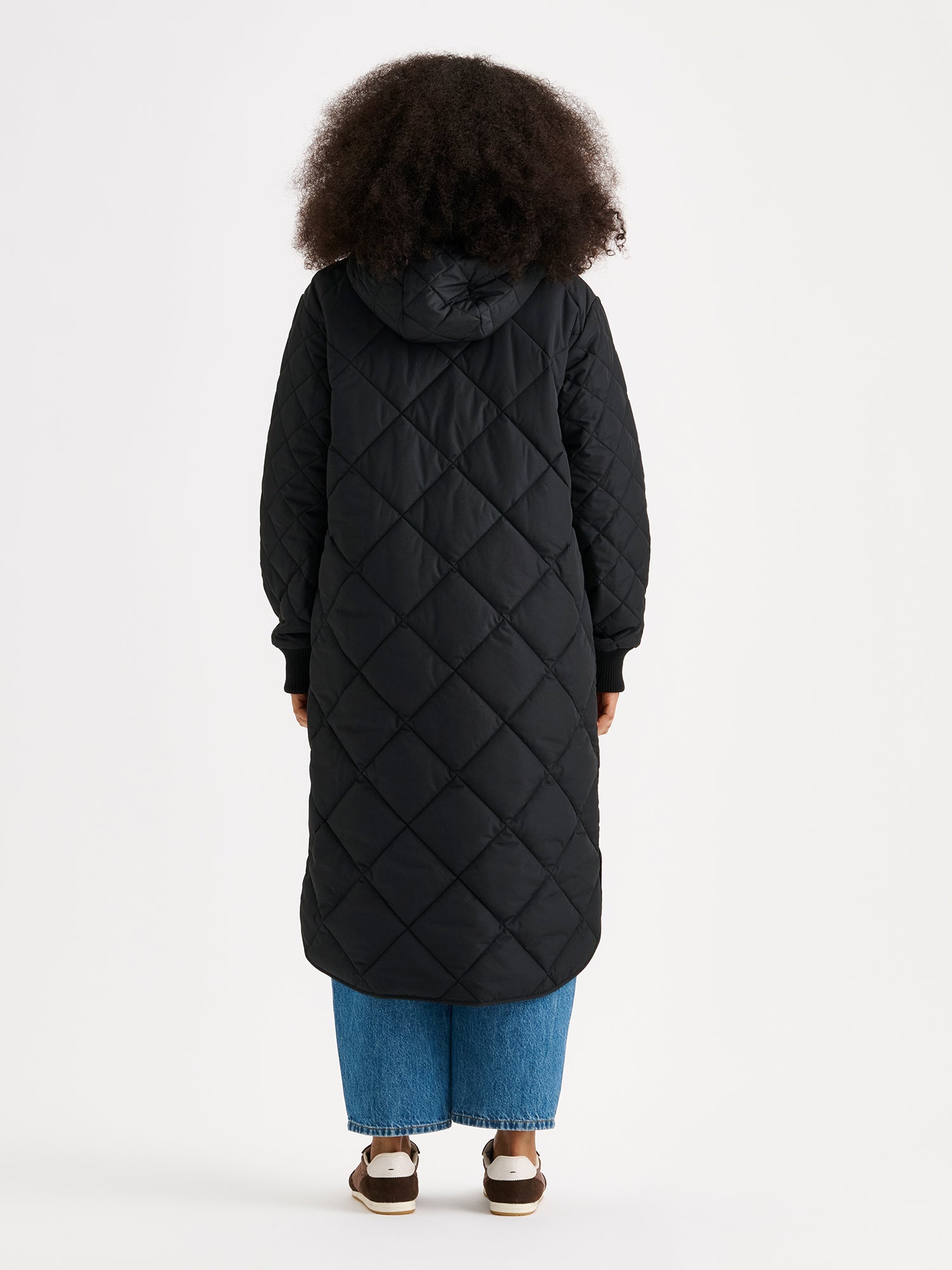 Nanso women's casual black Maxi quilted coat with light padding and a cozy hood. Perfect for in-between seasons, the hooded jacket features a two-way front zipper with a snap-button wind flap, a comfortable ribbed hood edge and cuffs. On a model photographed from the back side.