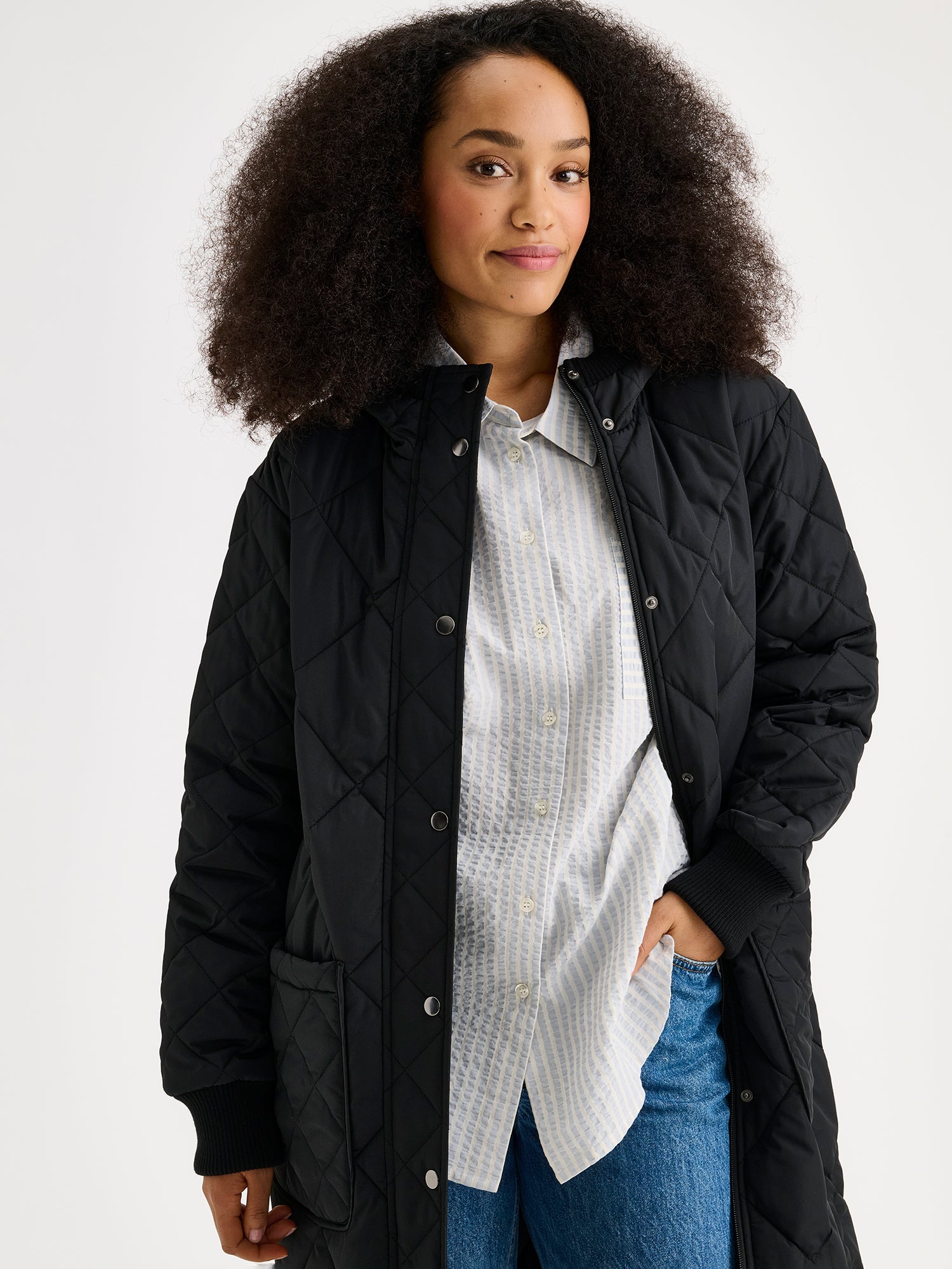Nanso women's casual black Maxi quilted coat with light padding and a cozy hood. Perfect for in-between seasons, the hooded jacket features a two-way front zipper with a snap-button wind flap, a comfortable ribbed hood edge and cuffs. On a model, close-up.